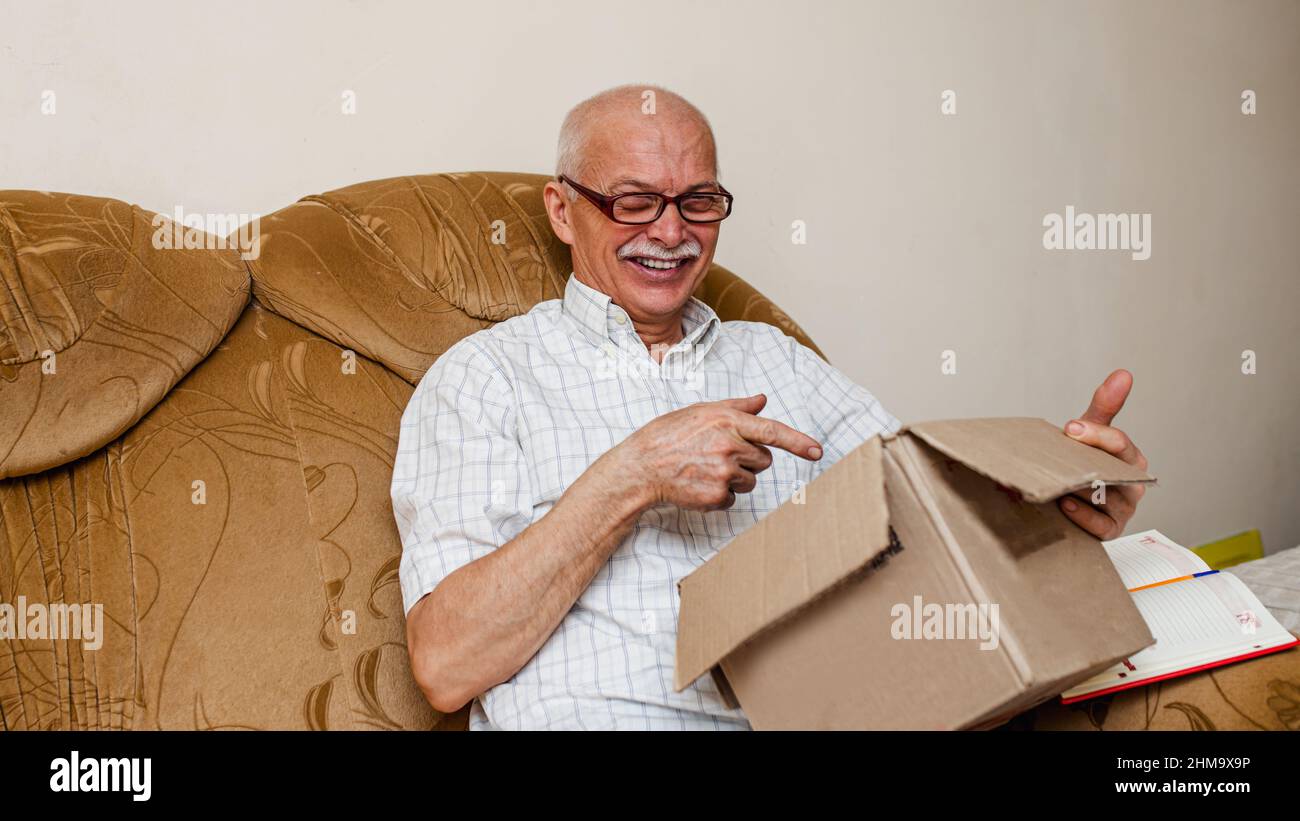 El anciano feliz maduro inspecciona el paquete recibido. El anciano se  sienta en un sofá vintage en la sala de estar. Pedido en línea para entrega  rápida de mensajería para pensionistas Fotografía