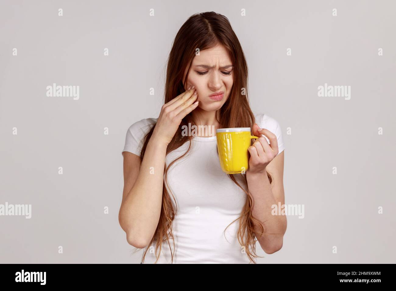 Mujer enferma malsana tocando el mentón sufriendo dolor de dientes, tiene dientes sensibles, siente dolor después de beber agua fría, usando una camiseta blanca. Estudio en interior grabado aislado sobre fondo gris. Foto de stock