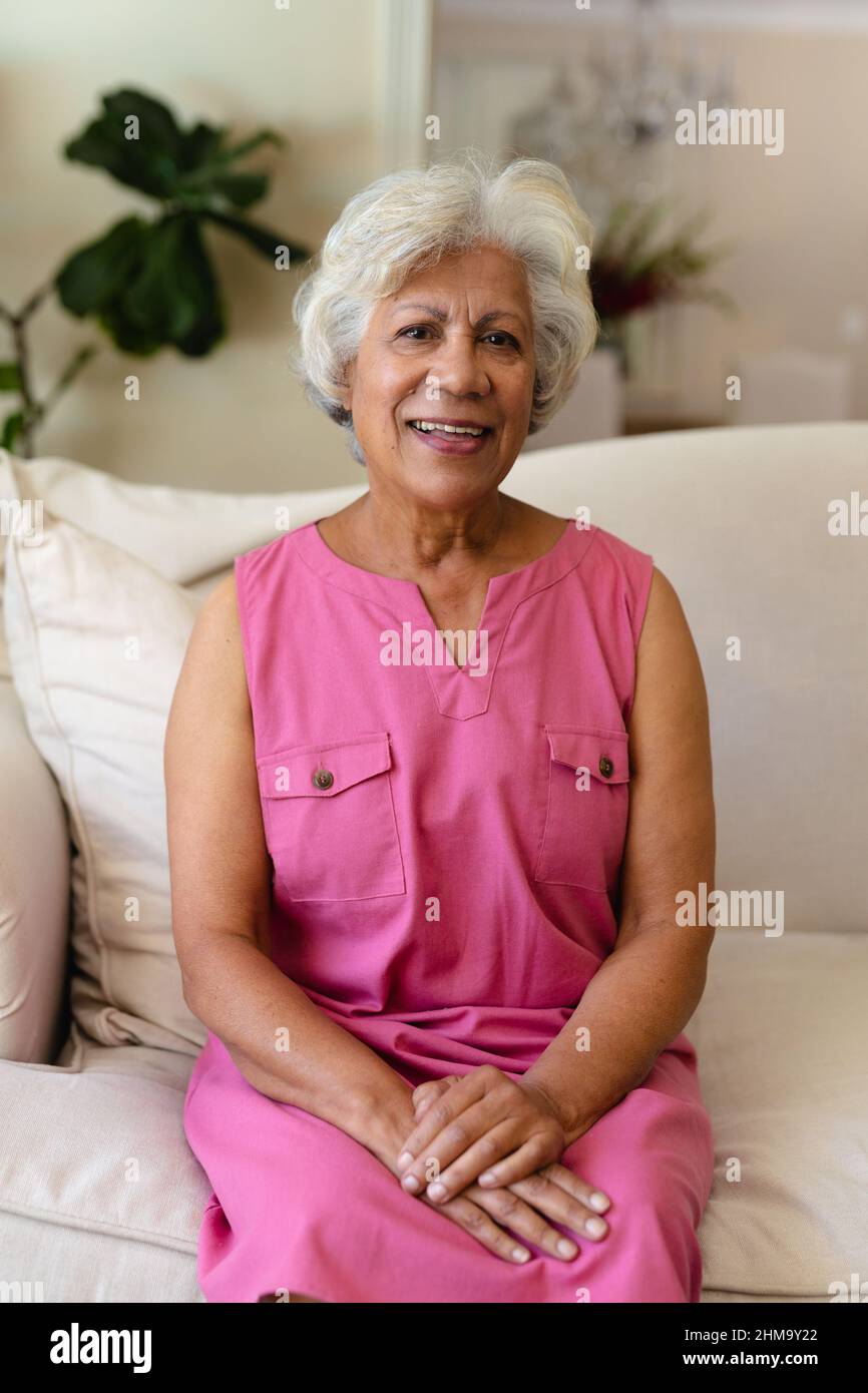 Retrato de una mujer madura biracial sonriente con pelo gris sentado en el sofá de la casa Foto de stock