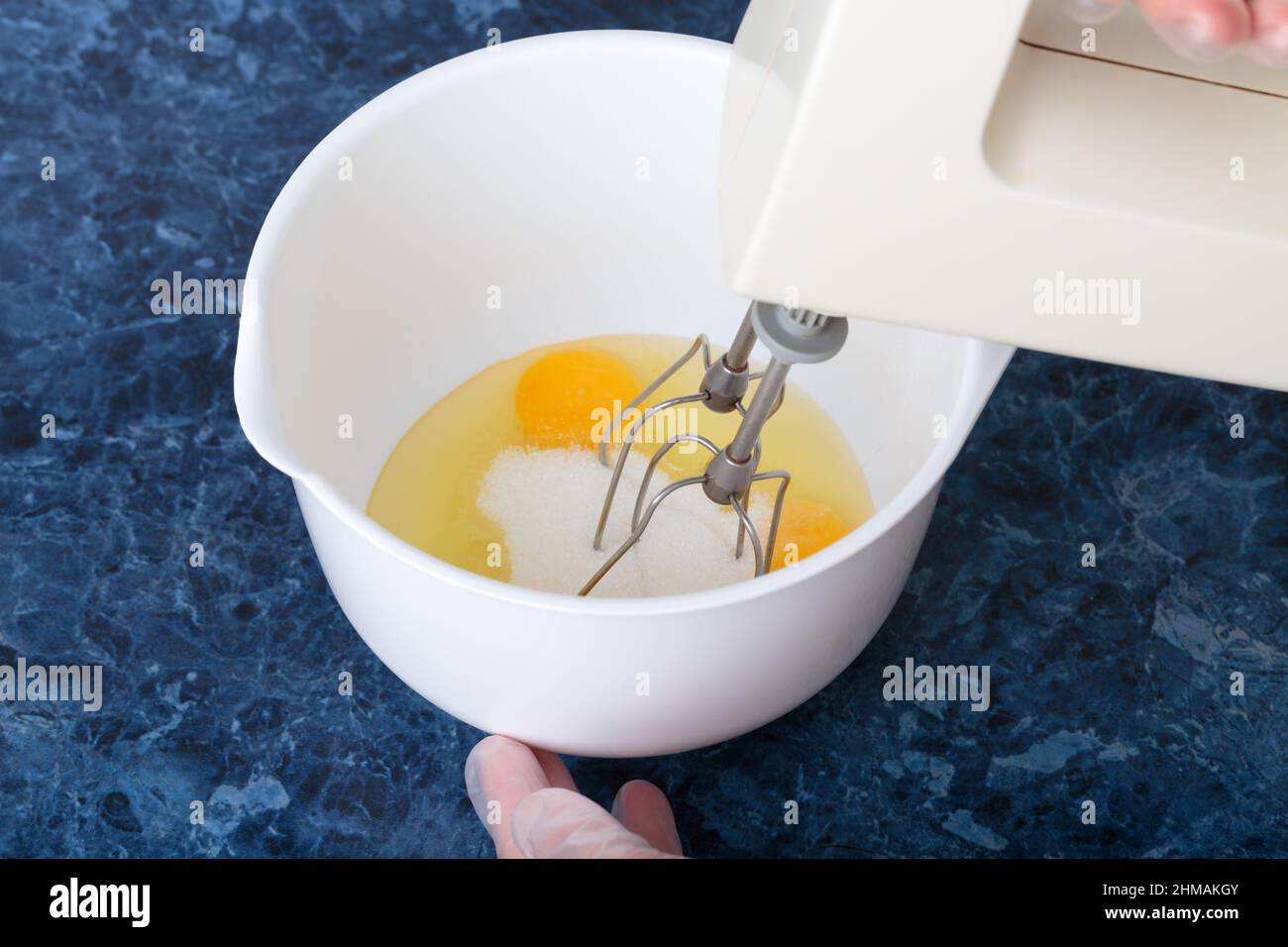 Una licuadora bate huevos y azúcar en un recipiente. Este proceso tiene  lugar en un tazón blanco. Fondo de mármol azul Fotografía de stock - Alamy