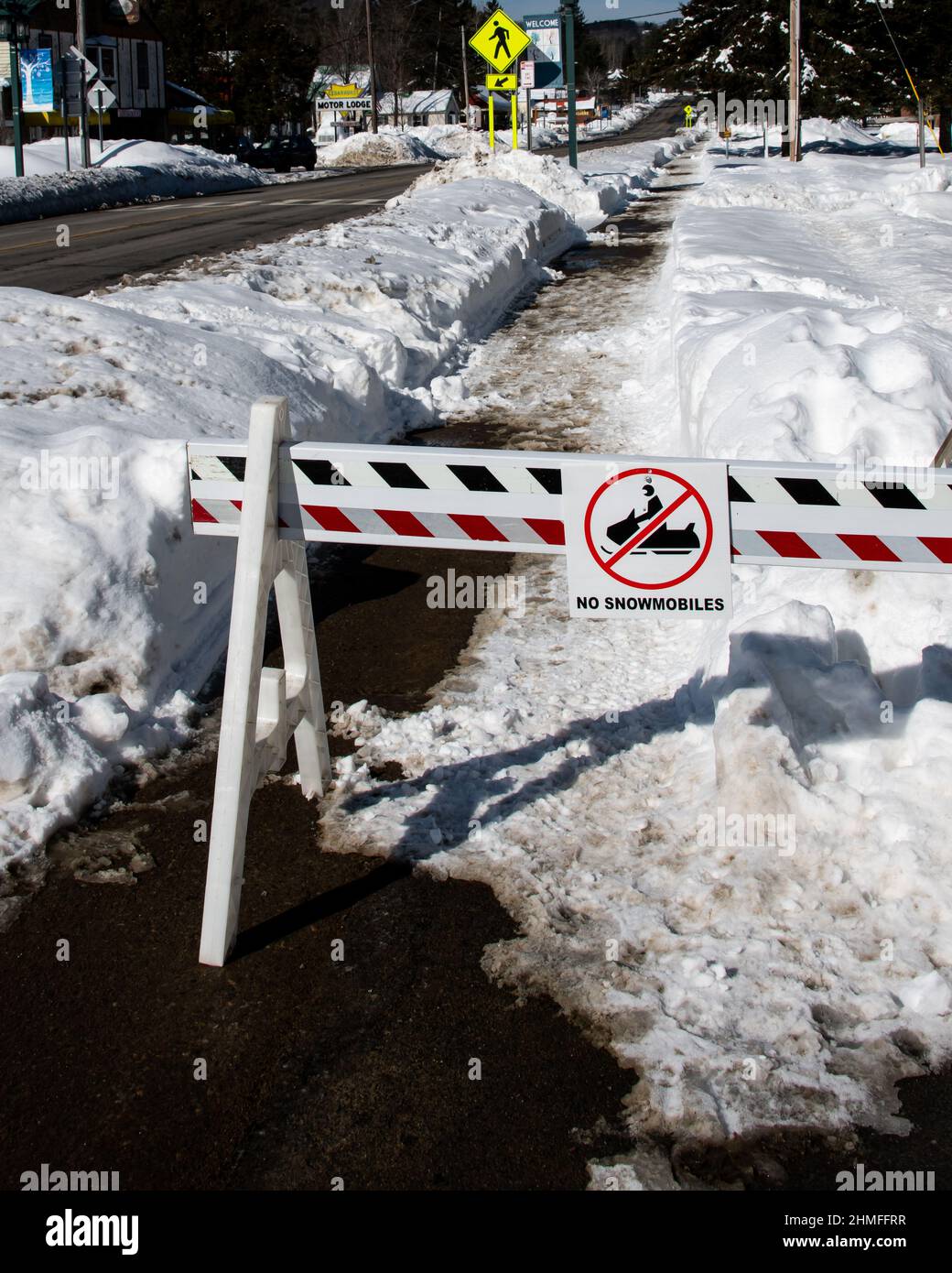 Una barricada roja y blanca y un cartel que indica que no se permiten motos de nieve en la acera con bancos de nieve profundos y hielo. Foto de stock