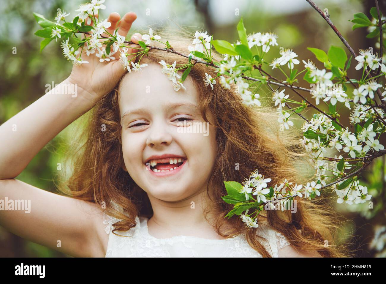 Riendo niña sin dientes vestido blanco cerca de árboles de cerezo florecientes. Sonrisa feliz y saludable. Foto de stock