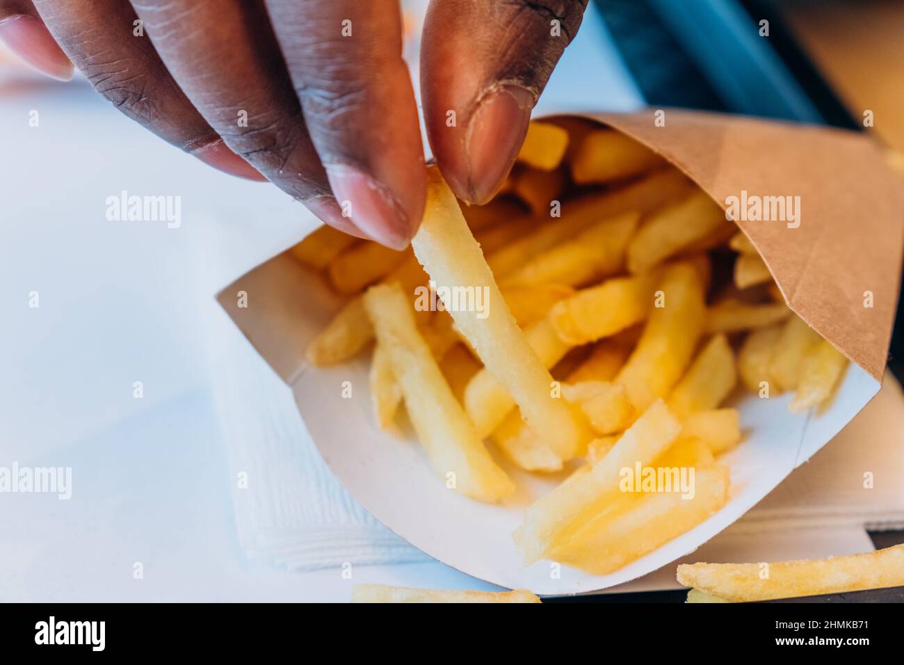 El joven afroamericano toma deliciosas patatas fritas frescas del recipiente de papel en la mesa en una moderna cafetería con vista muy cercana Foto de stock