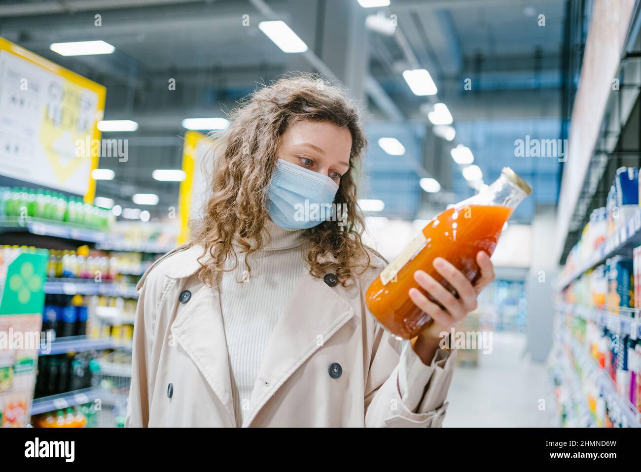 Una joven cuajada con una máscara médica y un impermeable beige escoge el jugo en una botella de vidrio en un supermercado, cerca Foto de stock