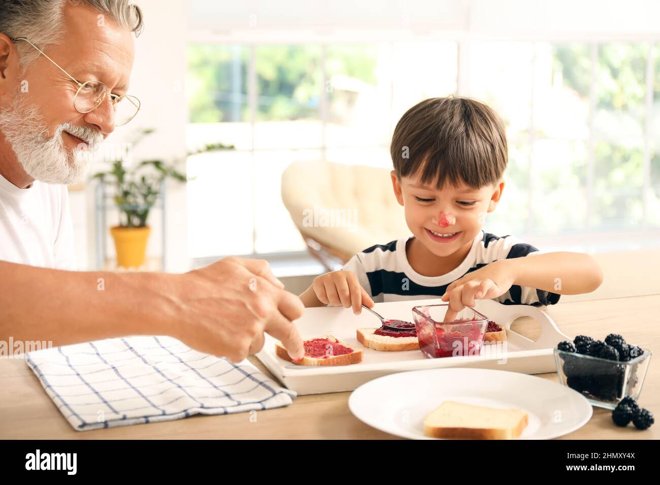 Niño pequeño y su abuelo haciendo sándwiches con mermelada en la mesa en la cocina Foto de stock