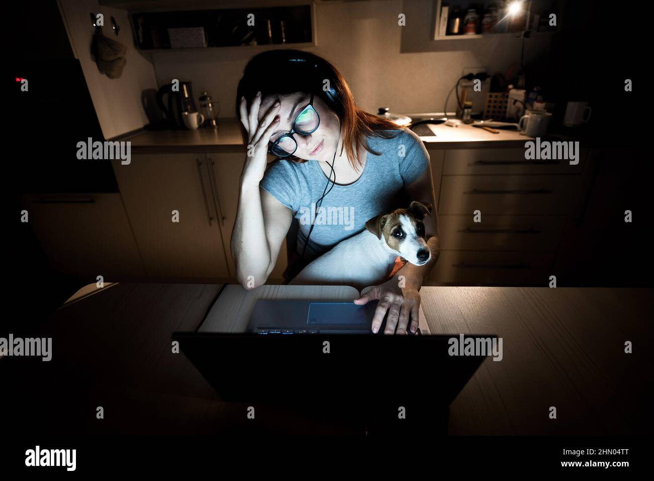 Mujer cansada con auriculares y gafas estudiando en un ordenador portátil. Una estudiante se prepara para un examen en la cocina tarde por la noche. Chica con un Foto de stock