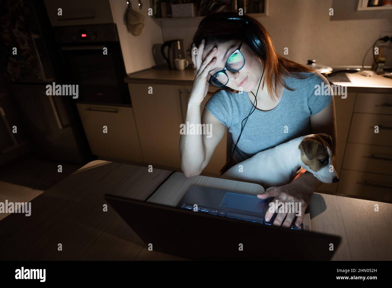 Mujer cansada con auriculares y gafas trabajando horas extras sobre un portátil. Mujer independiente trabajando a distancia en casa en la cocina tarde en la noche Foto de stock