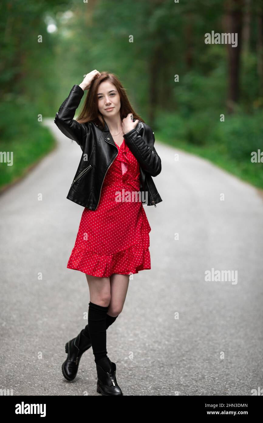 Una chica con un vestido rojo claro y aireado, una chaqueta de cuero en  estilo rocoso en una carretera vacía en un bosque de verano. Fotografías de  alta calidad Fotografía de stock -
