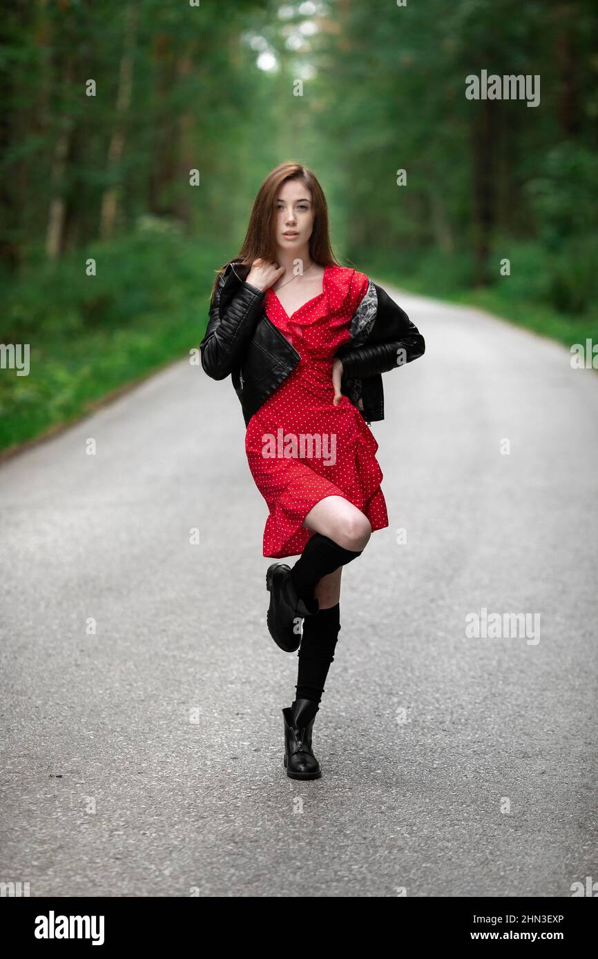 Una chica con un vestido rojo claro y aireado, una chaqueta de cuero en estilo rocoso en una carretera vacía en un bosque de verano Fotografía de stock - Alamy Una chica con un vestido rojo claro y aireado, una chaqueta de cuero en estilo rocoso en una carretera vacía en un bosque de verano Fotografía de stock - Alamy