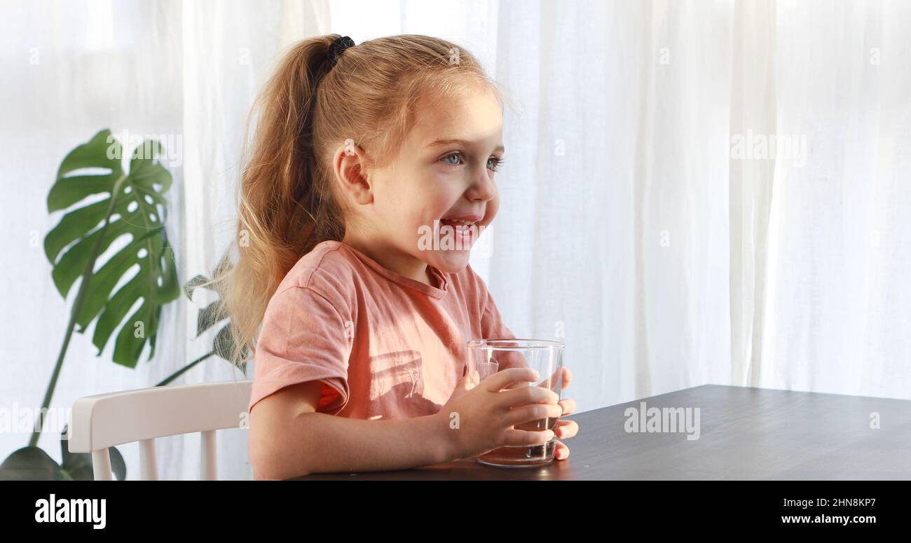 La niña rubia en vestido blanco bebe agua de un vaso en el interior día soleado. Un niño lindo está bebiendo una taza de agua Foto de stock