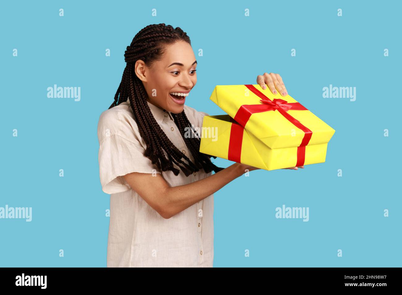 Retrato de feliz mujer emocionado con dreadlocks negro mirando en la caja de regalo, abrir el presente y mirar dentro con felicidad, llevando camisa blanca. Estudio en interior grabado aislado sobre fondo azul. Foto de stock