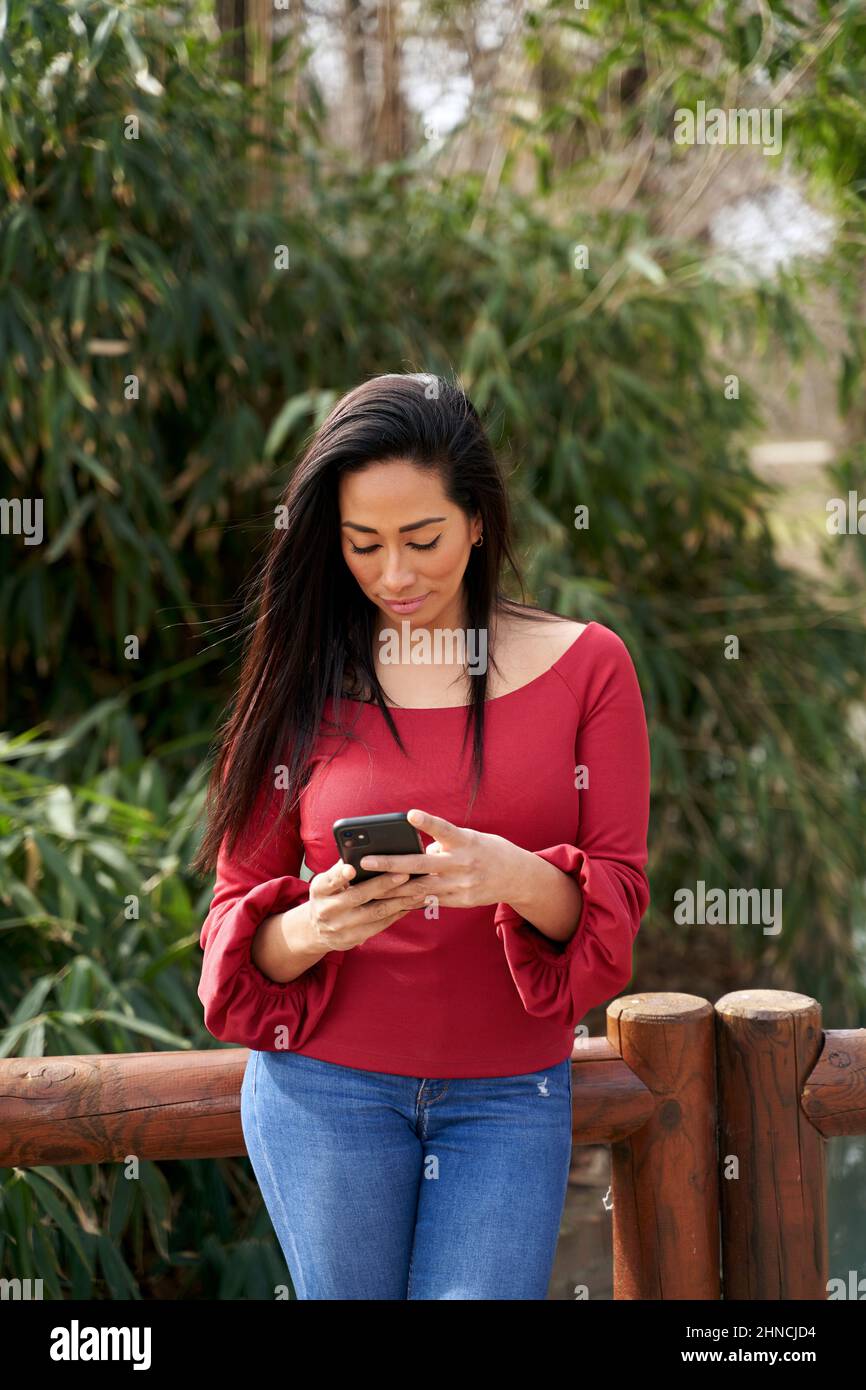 Hermoso mensaje de texto femenino hispano en el teléfono móvil mientras se encuentra cerca de la barandilla de madera en el parque con árboles verdes en el día de verano Foto de stock