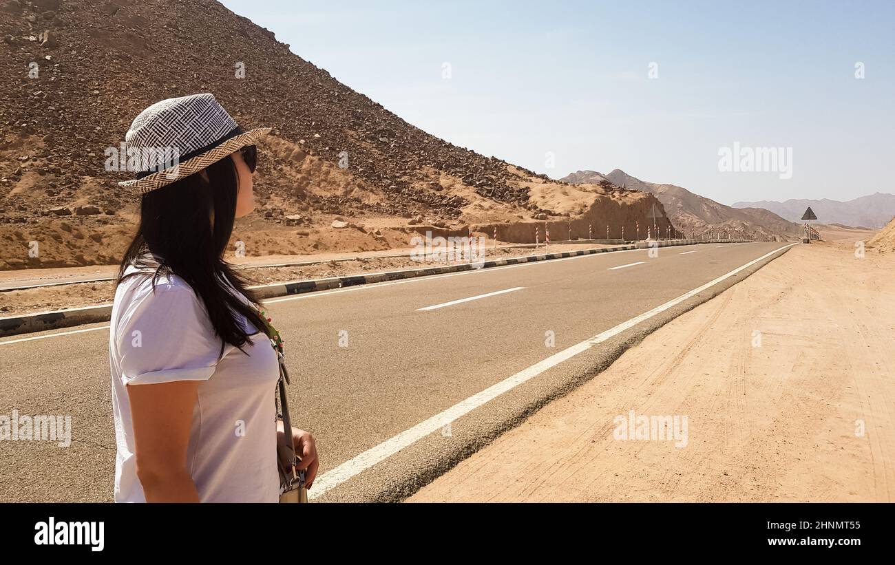 Una chica blanca en un sombrero se encuentra cerca de una amplia carretera de asfalto desierta. Retrato trasero de una mujer mirando a la distancia en un camino vacío con montañas y sol caliente. Cerca del este. Concepto de viaje Foto de stock