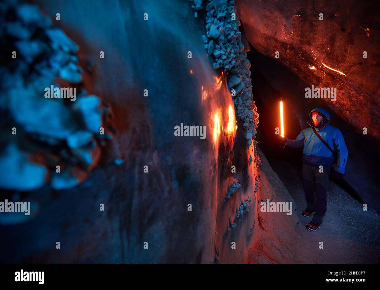 Un hombre de turismo que sostiene una lámpara incandescente roja en una cueva de hielo glacial profunda y estrecha con luz azul explora el glaciar de invierno de montaña en Kazajstán Foto de stock