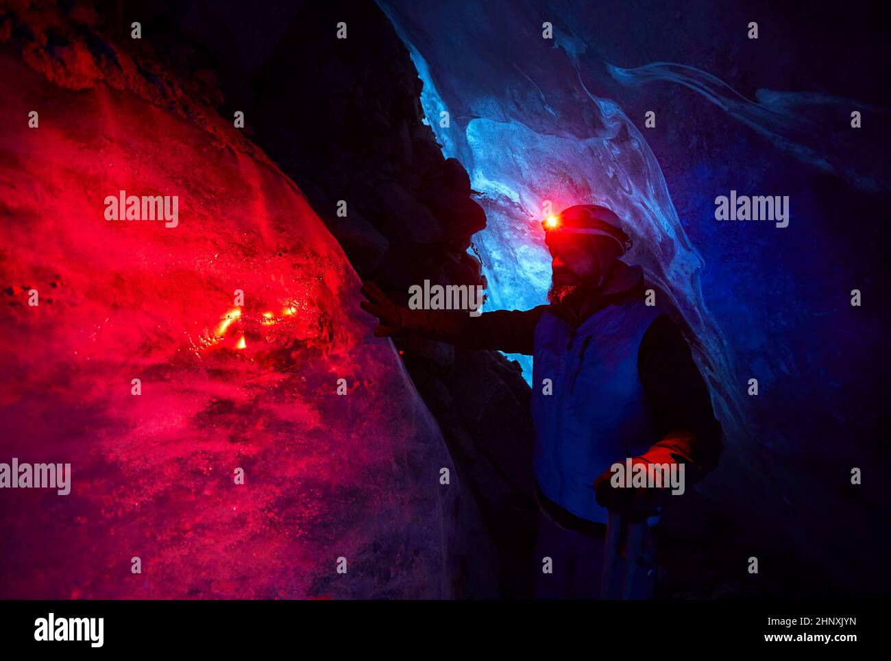 Alpinista con barba en casco con faro rojo brillante en profunda cueva de hielo glacial con luces azules explorar glaciar de montaña de invierno en Kazajstán Foto de stock
