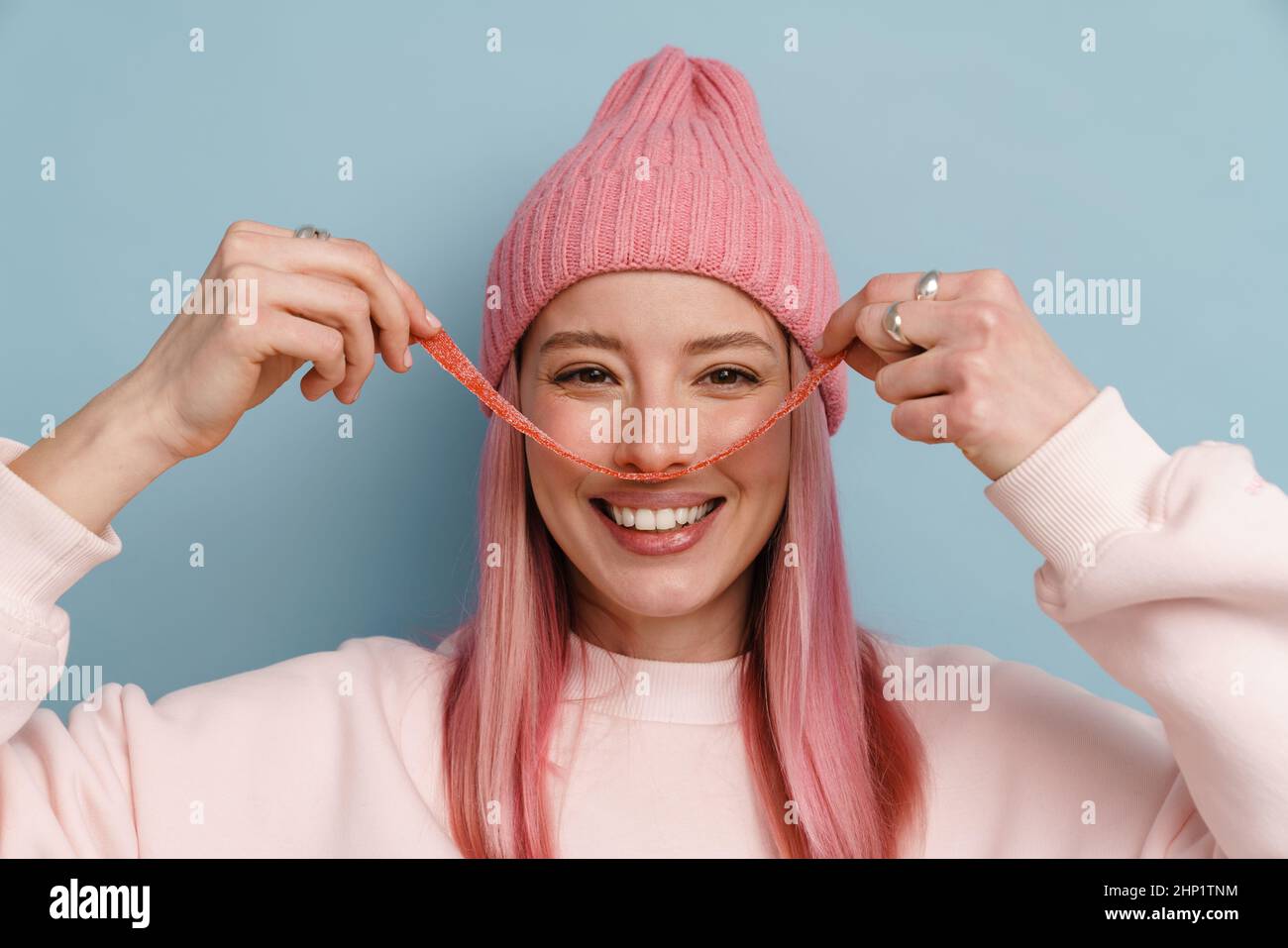 Mujer blanca joven con el pelo rosado sonriendo mientras sostenía el caramelo de la tira aislado sobre la pared azul Foto de stock