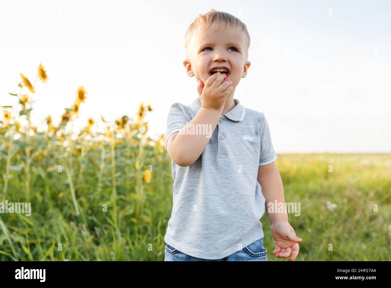 Niño pequeño jugando en el fondo del campo y sonriendo. Tres años de diversión en el campo de girasoles al atardecer. Estilo de vida al aire libre en verano Foto de stock