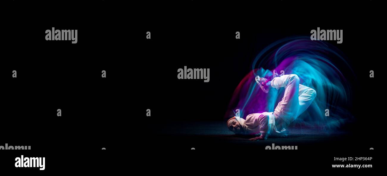 Un joven deportivo flexible y energético bailando hip-hop o breakdance en traje blanco sobre fondo oscuro con una mezcla de luces de neón azules. Deporte, arte, acción Foto de stock