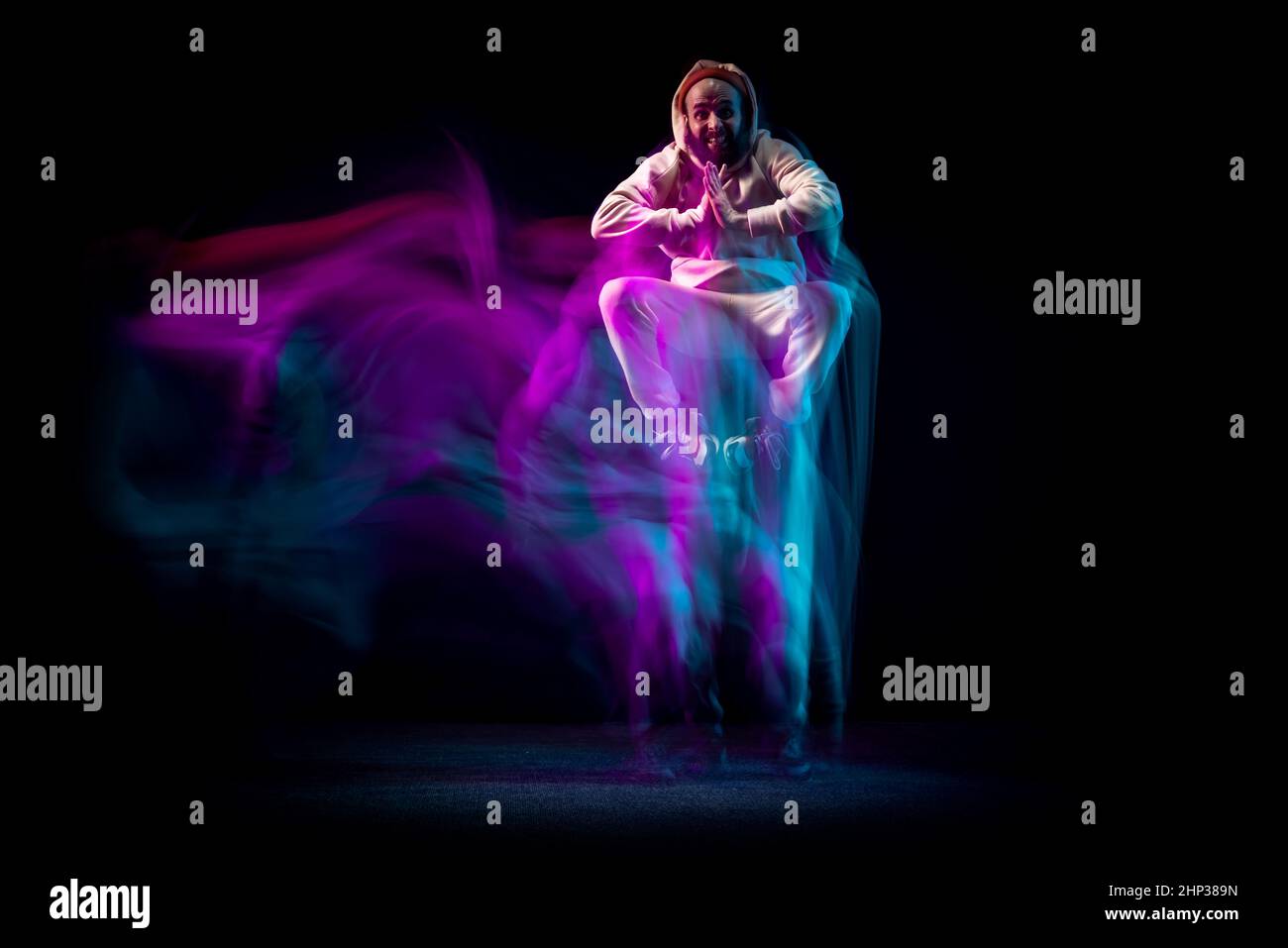 Un joven deportivo flexible y energético bailando hip-hop o breakdance en traje blanco sobre fondo oscuro con una mezcla de luces de neón azules. Deporte, arte, acción Foto de stock