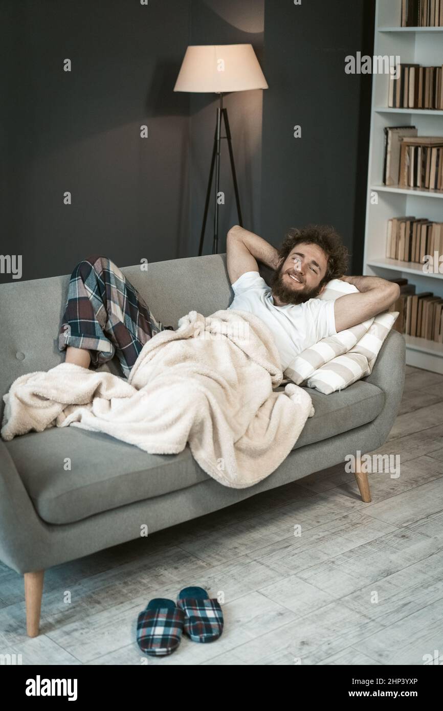 Retrato de un hombre relajado y guapo con pelo rizado durmiendo en el sofá o sofá, disfrutando de los días de fin de semana o de cuarentena en casa con una pequeña biblioteca en el fondo. Foto de stock