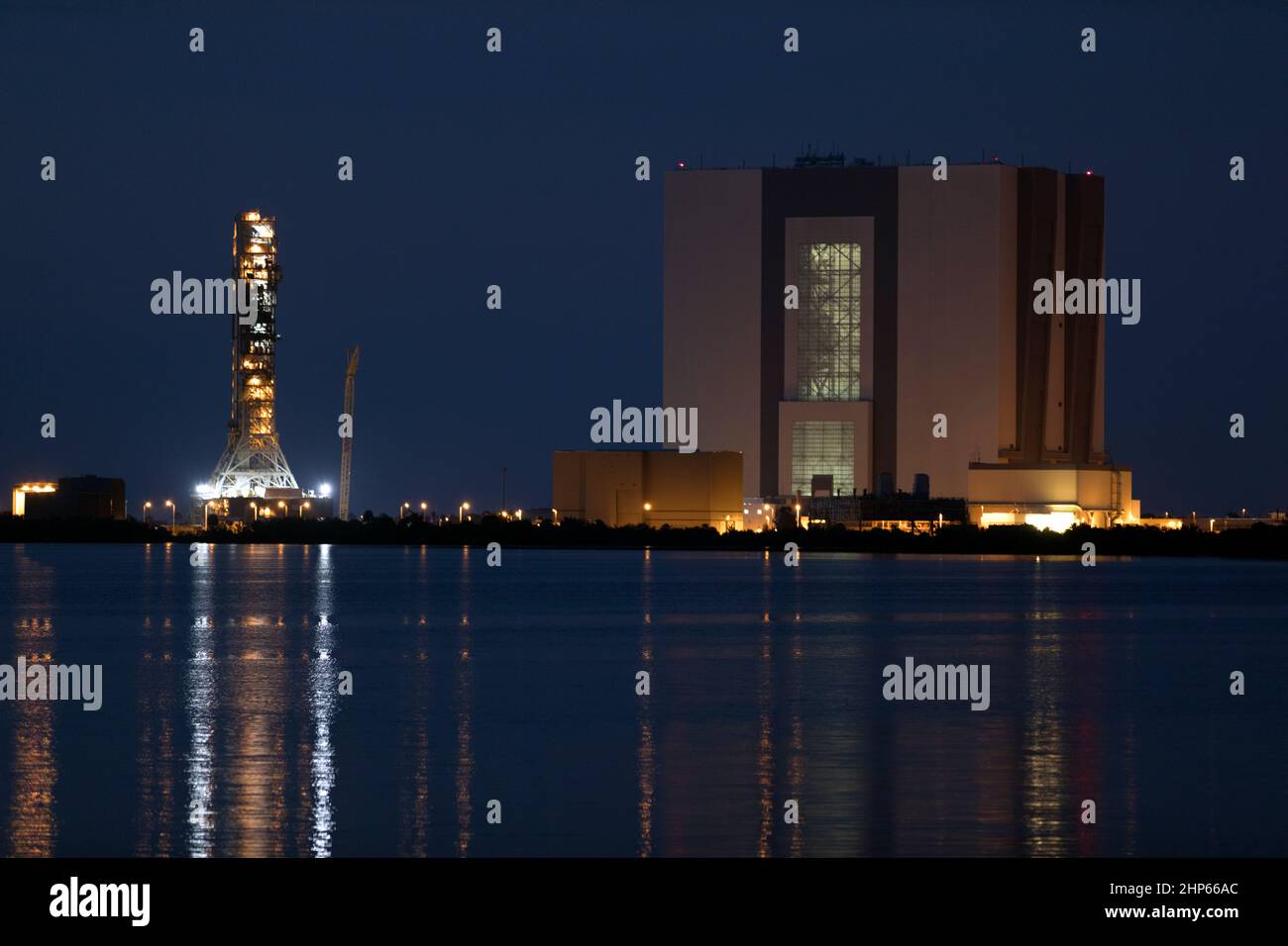 Con sus luces reflejadas en el agua, una vista nocturna del emblemático edificio de la Asamblea de Vehículos, a la derecha, y lanzador móvil en el Centro Espacial Kennedy de la NASA en Florida ca. 2018 Foto de stock