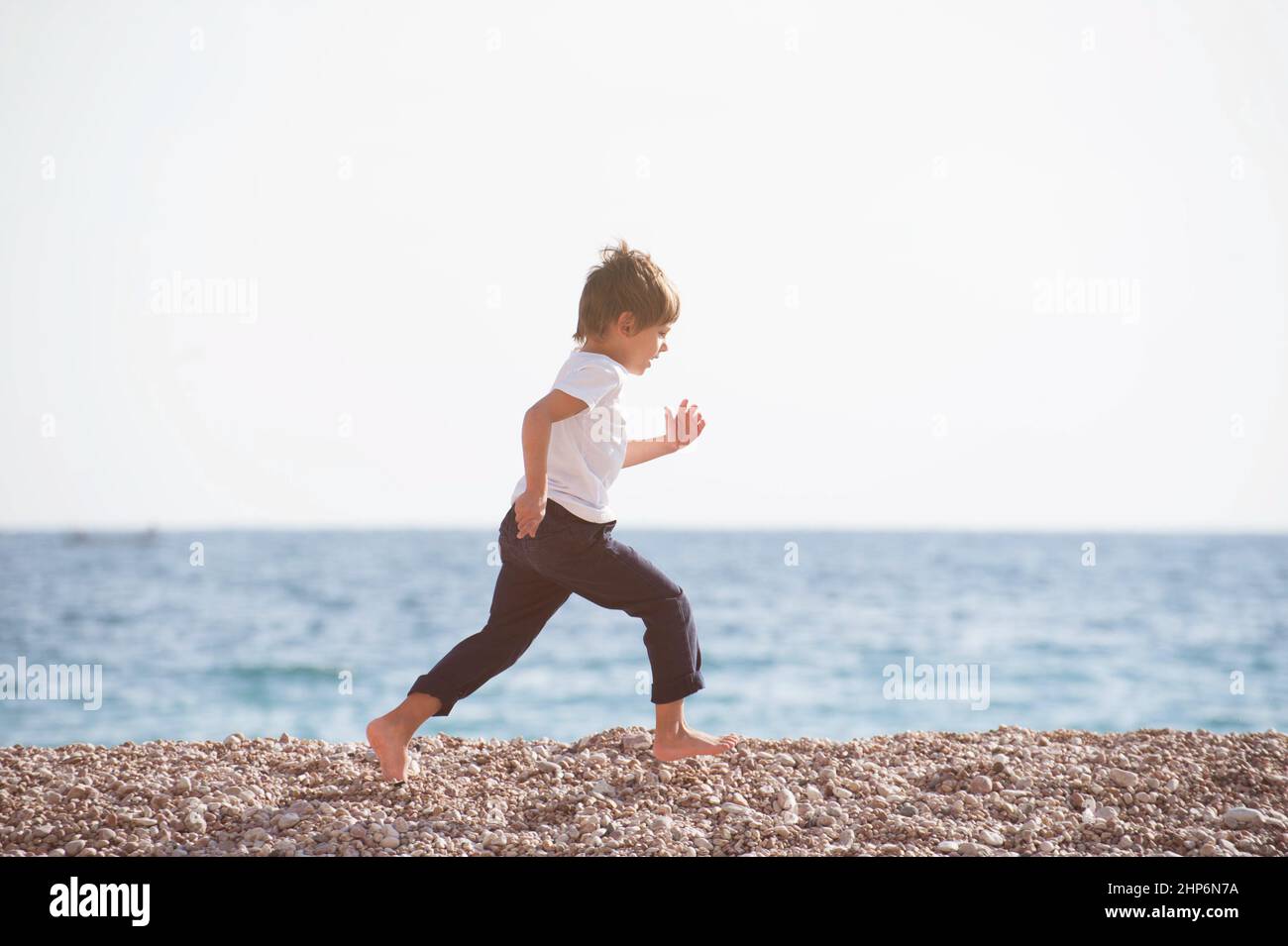 niño pequeño activo con jersey blanco y pantalones azules corriendo por la playa durante actividades deportivas al aire libre Foto de stock