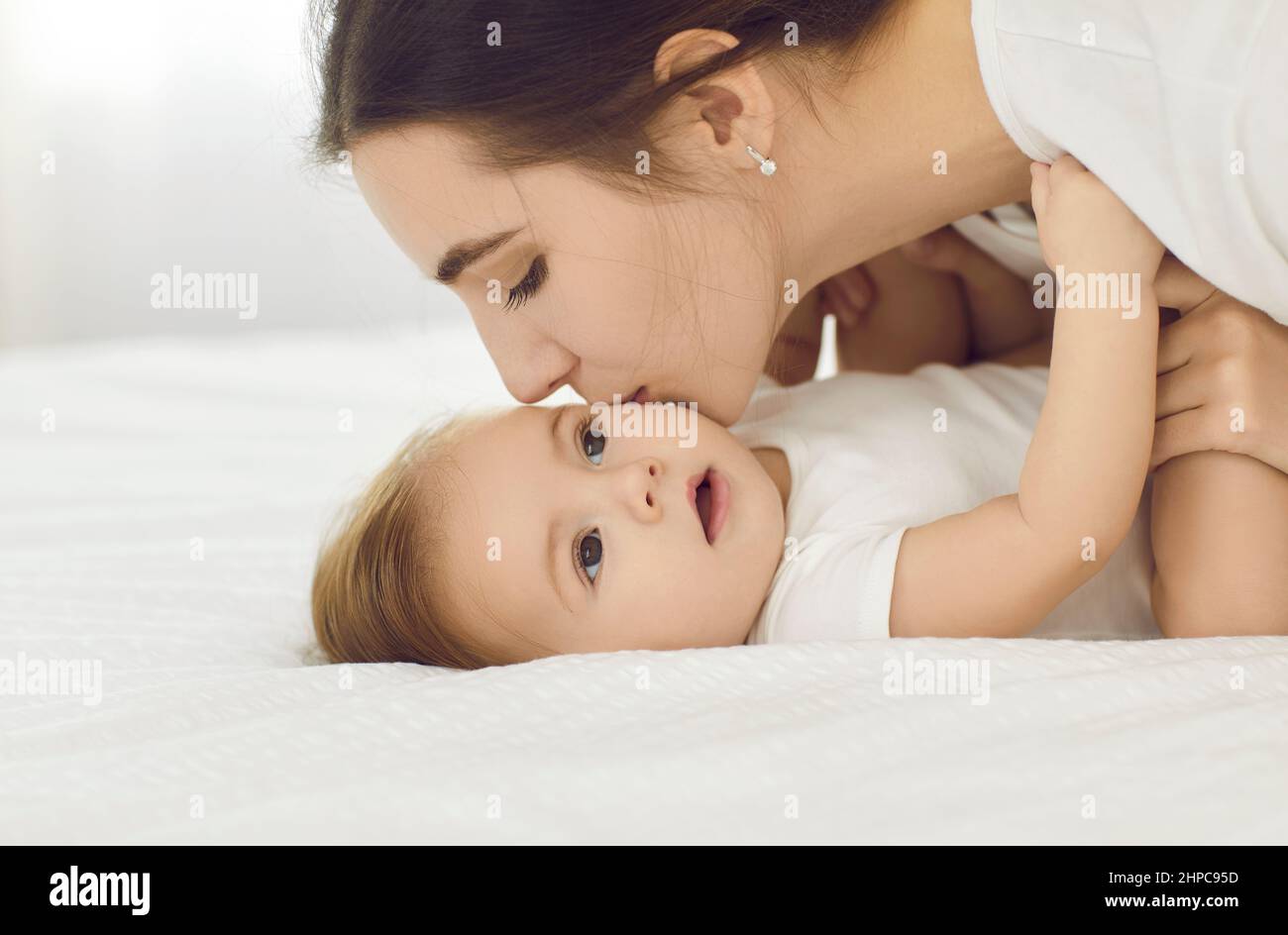 Feliz madre besando a su bebé niña en su pequeña mejilla suave mientras se cuddling en la cama juntos Foto de stock