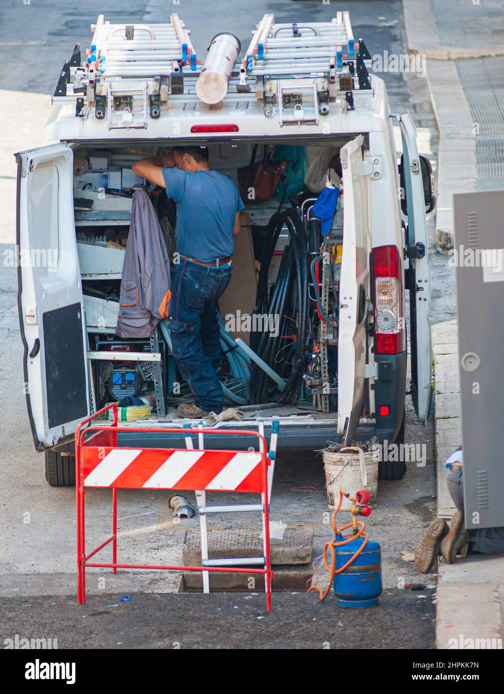 Trabajador dentro de su furgoneta, cargado con herramientas, protegido del tráfico con conos durante la reparación. Foto de stock