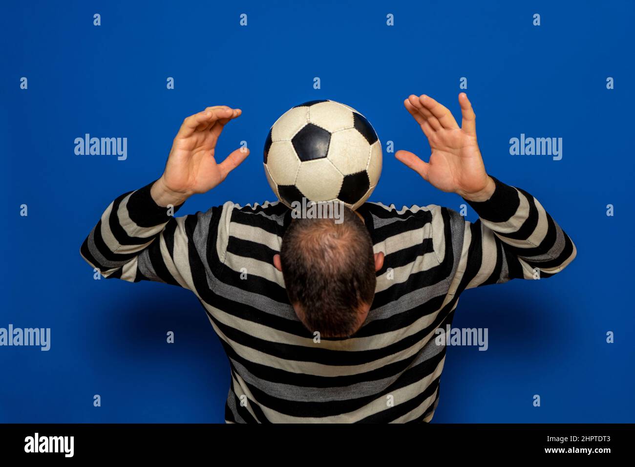 Hombre hispano con una calvicie incipiente tratando de hacer malabares a  una pelota de fútbol, tiene la pelota en la parte posterior de su cuello  mientras levanta sus brazos. Aislado sobre azul
