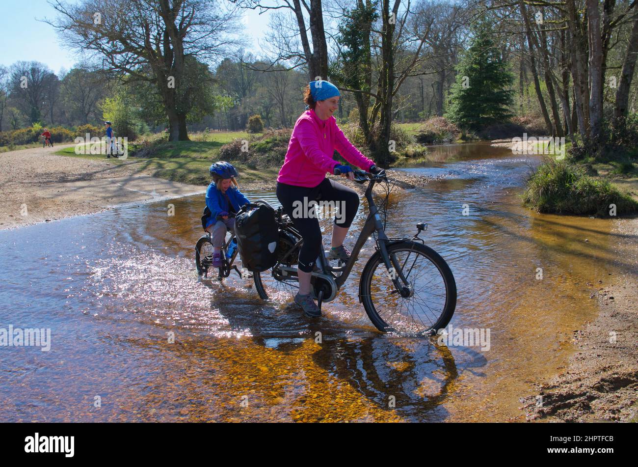Mujer en bicicleta eléctrica y niño en un tagalong (ciclo de remolque) a través de Fletchers Water ford en el Parque Nacional de New Forest/ Foto de stock