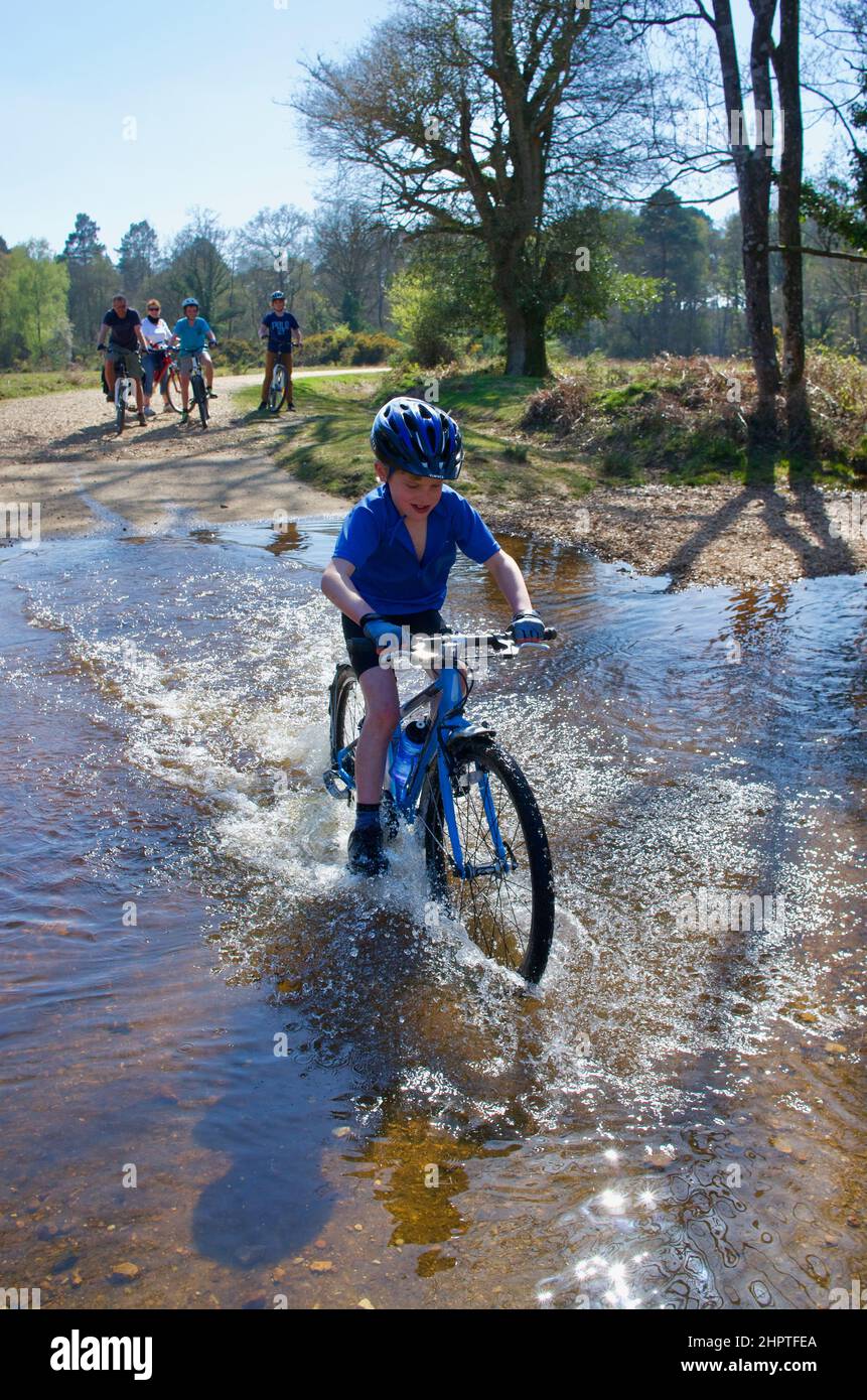 Un niño monta en bicicleta con confianza a través del Fletchers Water Ford en el Parque Nacional New Forest, con diversión y salpicaduras, como otros ciclistas miran. Foto de stock