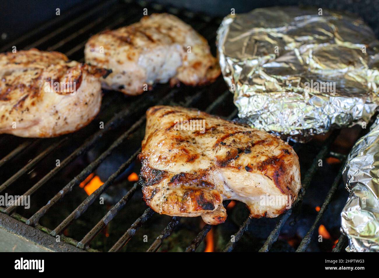 Pollo cocido en la parrilla: Pechugas de pollo deshuesadas asando en una parrilla con paquetes envueltos en papel de aluminio en el fondo. Foto de stock