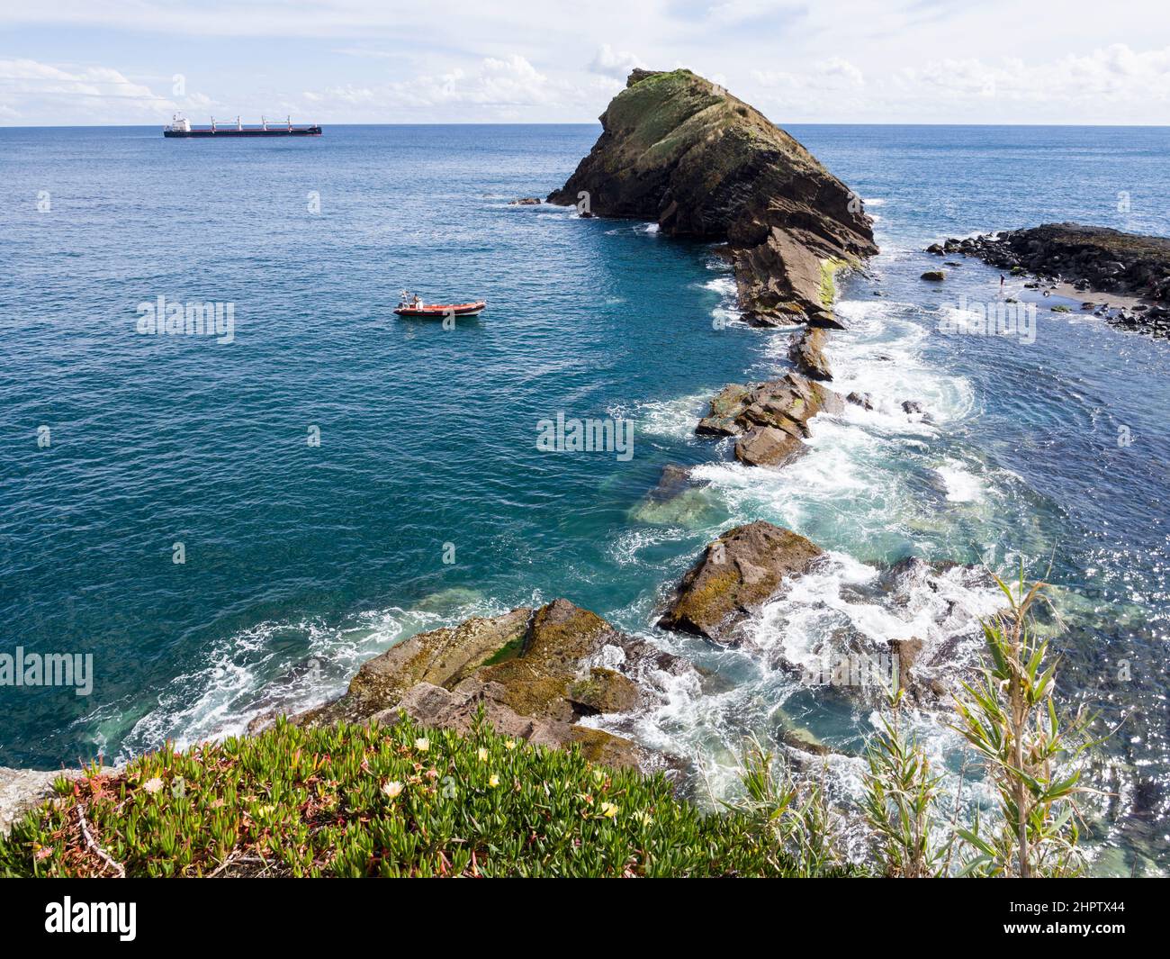 La Roca de Sao Roque: Una roca icónica se adentra en el Océano Atlántico. Un pequeño bote de pesca rojo y un gran carguero están amarrados cerca por un hombre que se desfila en una playa cercana. Foto de stock