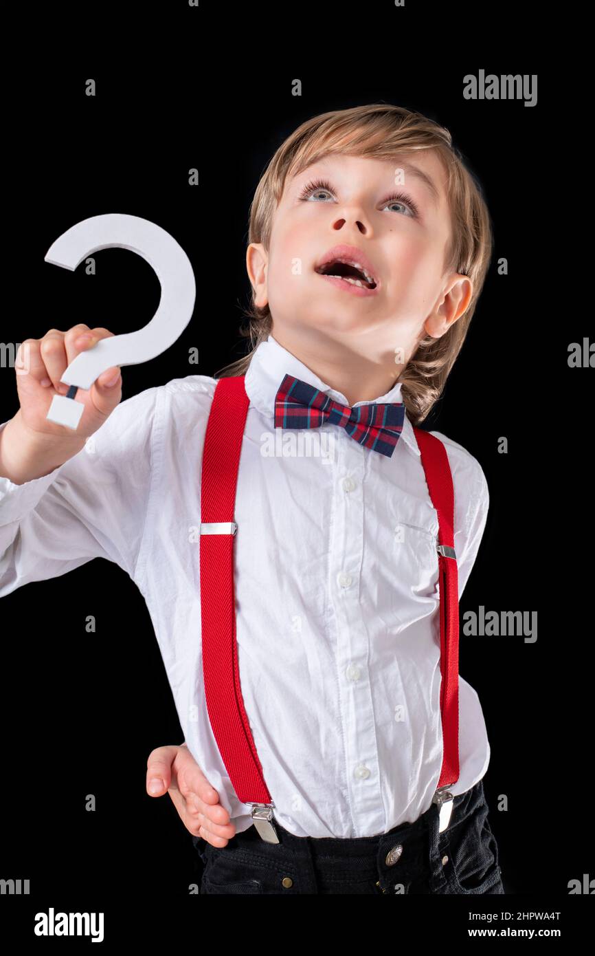 Adorable niño preescolar muy bien vestido sosteniendo un signo de pregunta sobre el fondo negro y mirando hacia arriba. Foto de stock