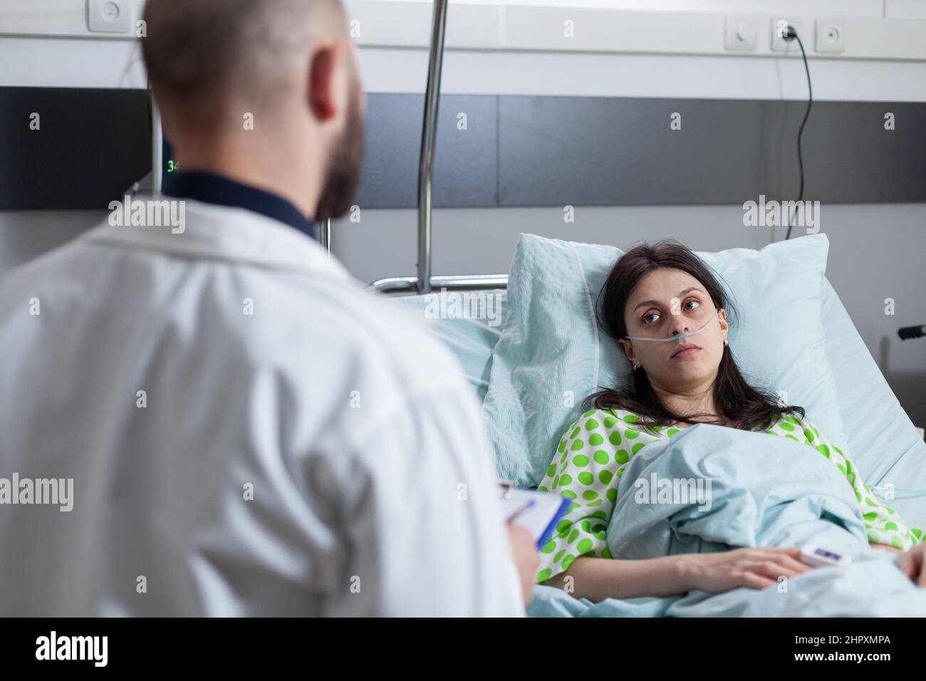 Vista sobre el hombro de una mujer que recibe oxígeno en la cama del hospital mirando al médico sosteniendo el portapapeles con resultados de laboratorio. Paciente con cánula nasal con baja saturación de SpO2 en sala de hospital. Foto de stock