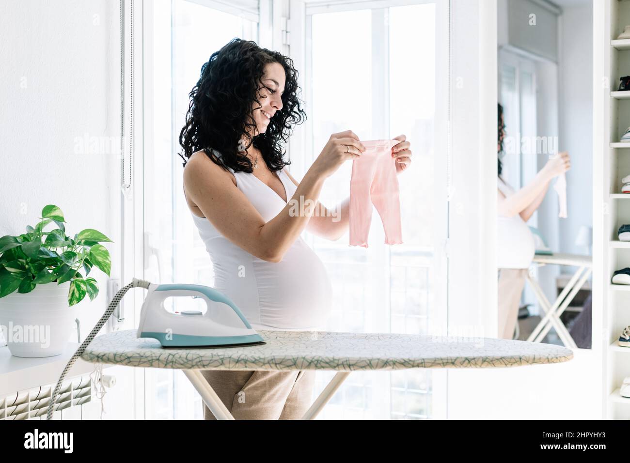 joven mujer embarazada bonita con pelo rizado plancha la ropa de su futuro bebé, preparando la habitación para el recién nacido Foto de stock