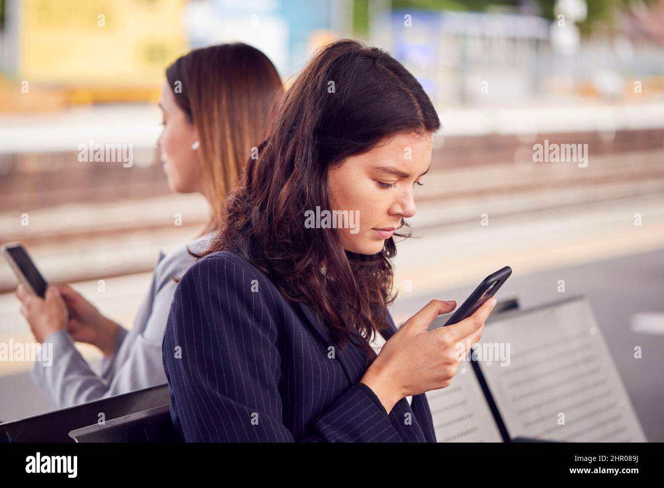 Dos mujeres de negocios que se desplazaban al trabajo esperando la plataforma de tren en la estación mirando los teléfonos móviles Foto de stock