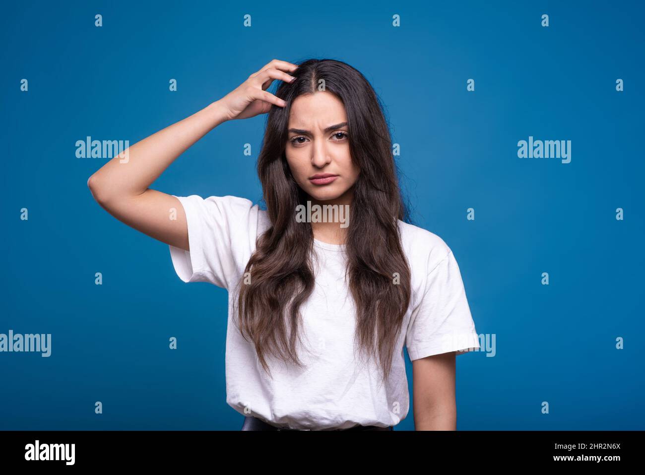 Atractiva mujer caucásica o morena árabe en una camiseta blanca que sostiene su mano en la cabeza y piensa aislada en un fondo de estudio azul. Foto de stock