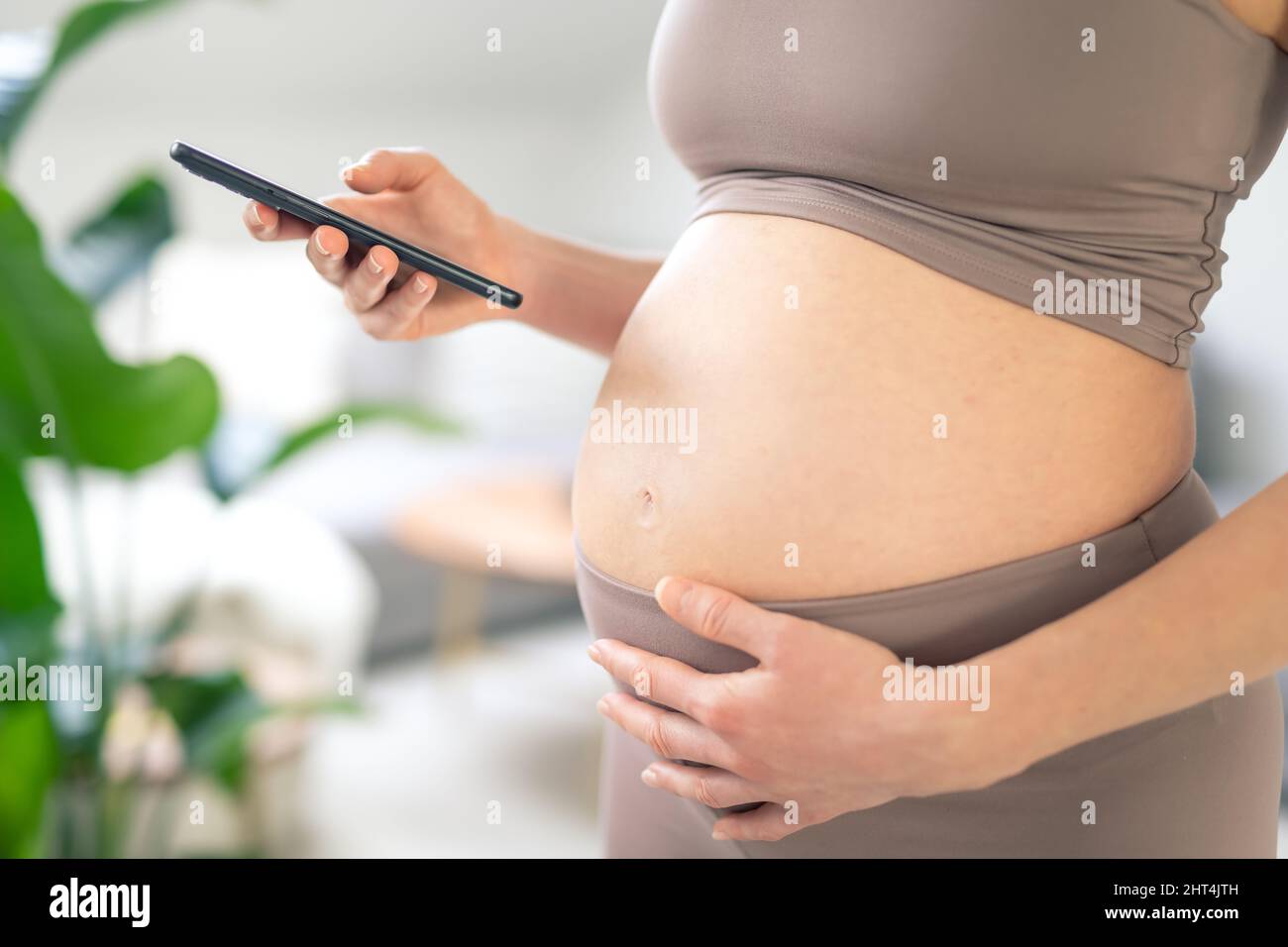Foto de cerca del vientre femenino embarazada. Mujer sosteniendo y utilizando la aplicación de teléfono inteligente móvil en el interior del hogar. Embarazo, tecnología, en línea Foto de stock