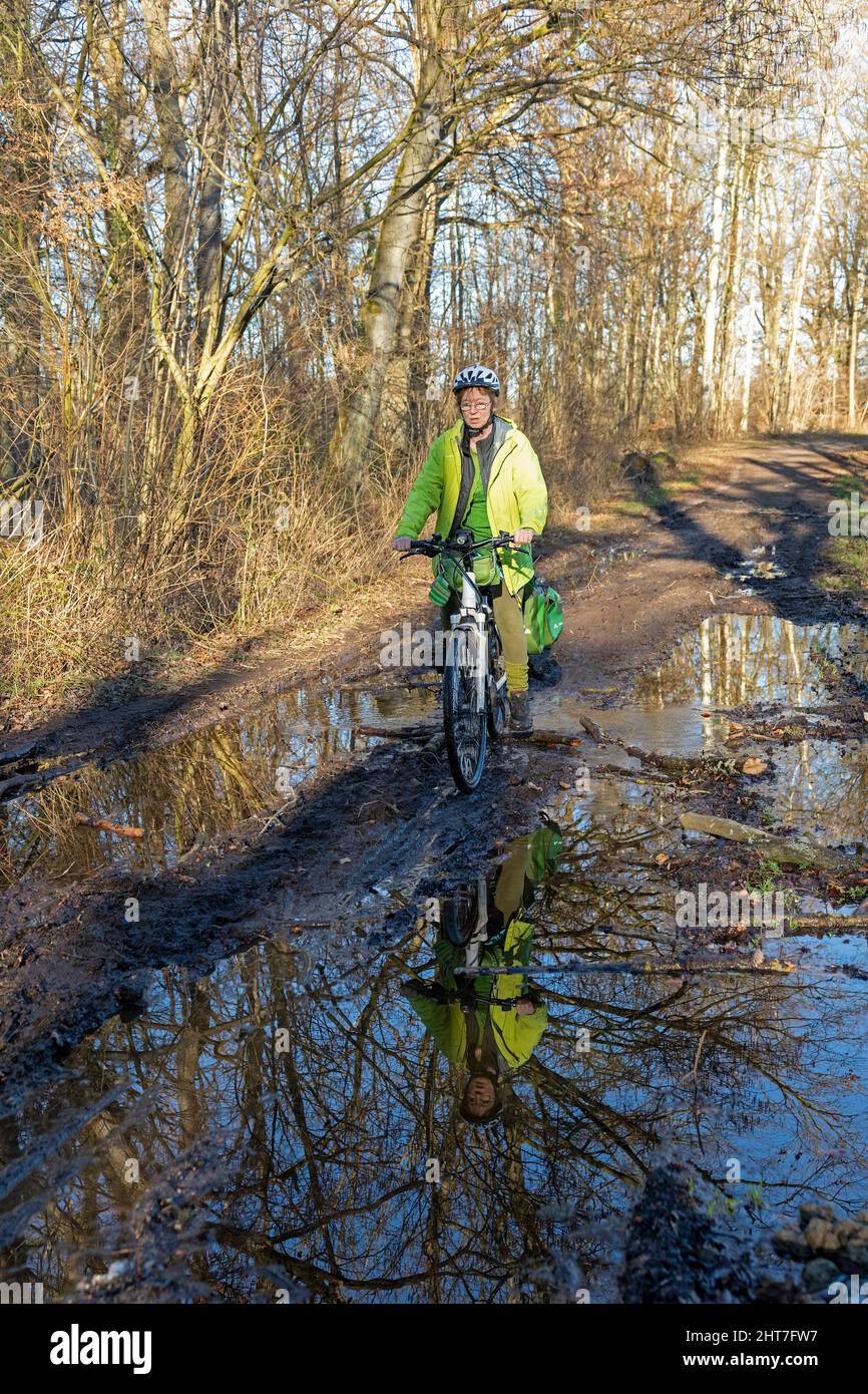 Mujer de más de cincuenta años haciendo un recorrido en bicicleta con su e-bike a lo largo de un camino fangoso y a través de charcos, Lueneburg, Baja Sajonia, Alemania Foto de stock
