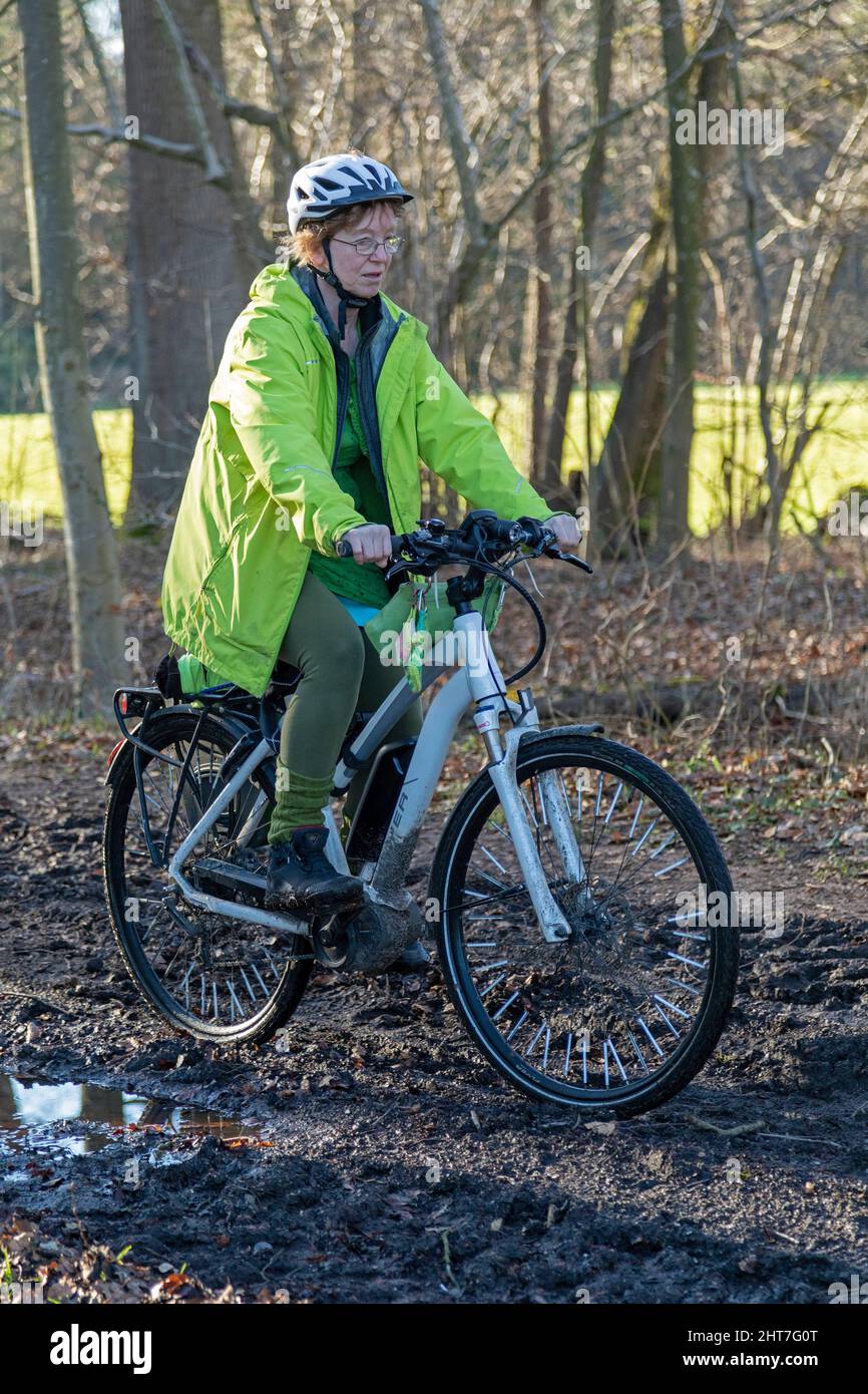 Mujer de más de cincuenta años haciendo un recorrido en bicicleta con su e-bike a lo largo de un camino fangoso y a través de charcos, Lueneburg, Baja Sajonia, Alemania Foto de stock