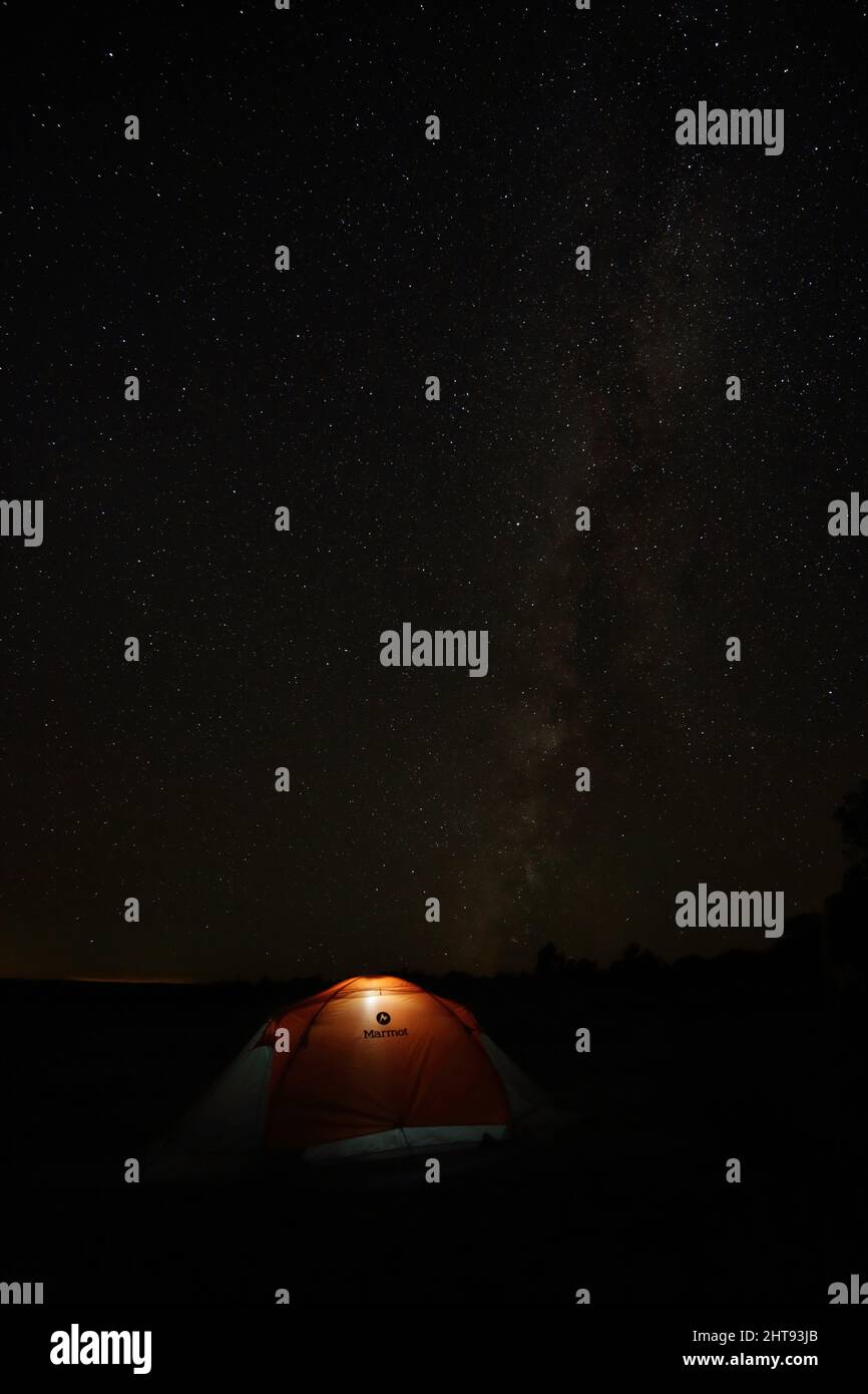 Vista de la Vía Láctea y el cielo nocturno con una tienda de campaña naranja iluminada en un campamento en el borde sur del Gran Cañón. En la distancia las luces de vinieron Foto de stock