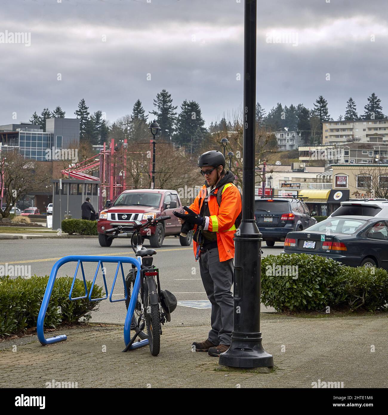Hombre en un chaleco de seguridad naranja poniendo sus guantes para montar su bicicleta que está en un soporte azul de bicicleta. Foto de stock