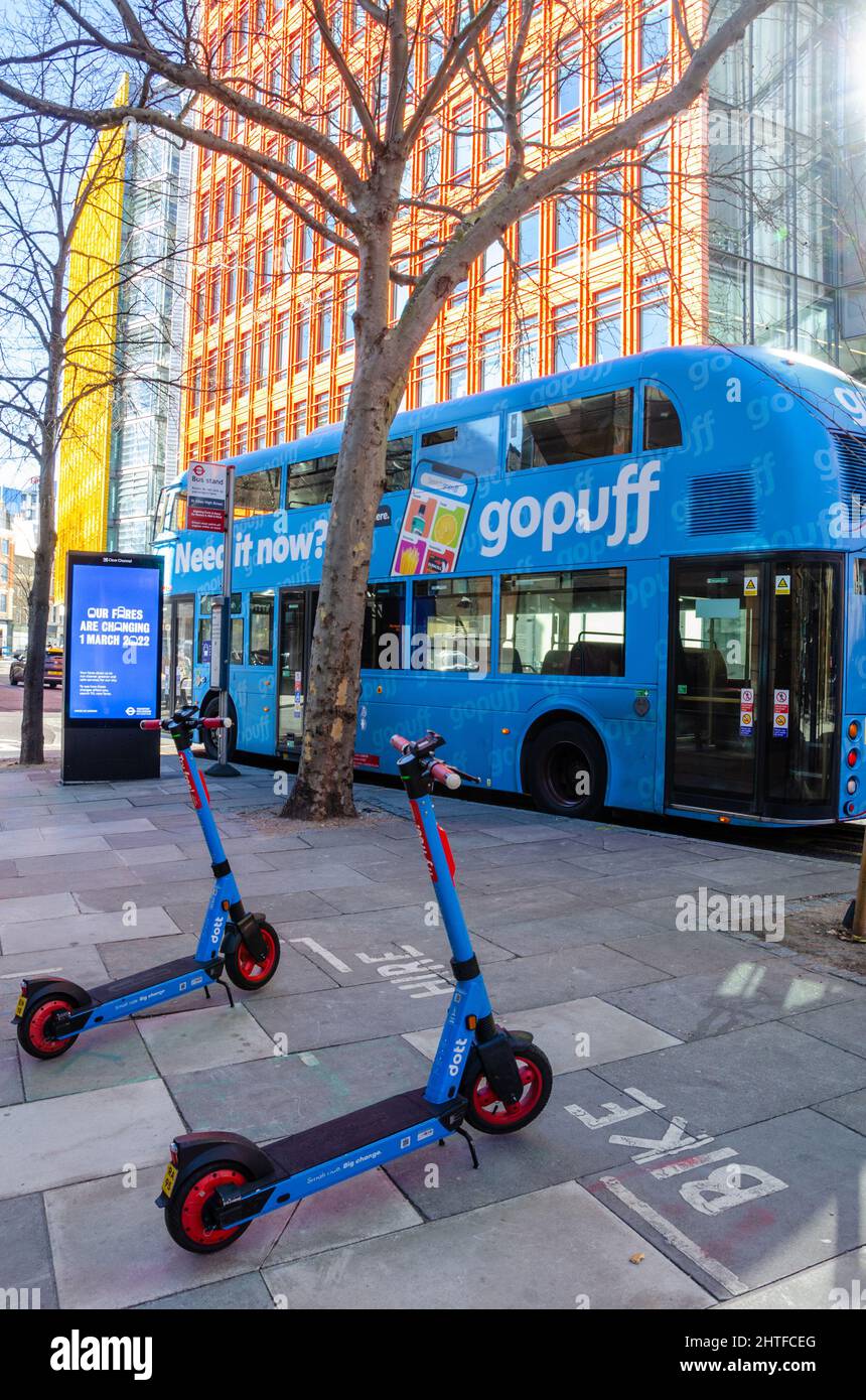 Scooters eléctricos de alquiler azules estacionados en un pavimento junto a un autobús azul de Londres frente a modernos edificios de oficinas en el centro de Saint Giles en Londres Foto de stock