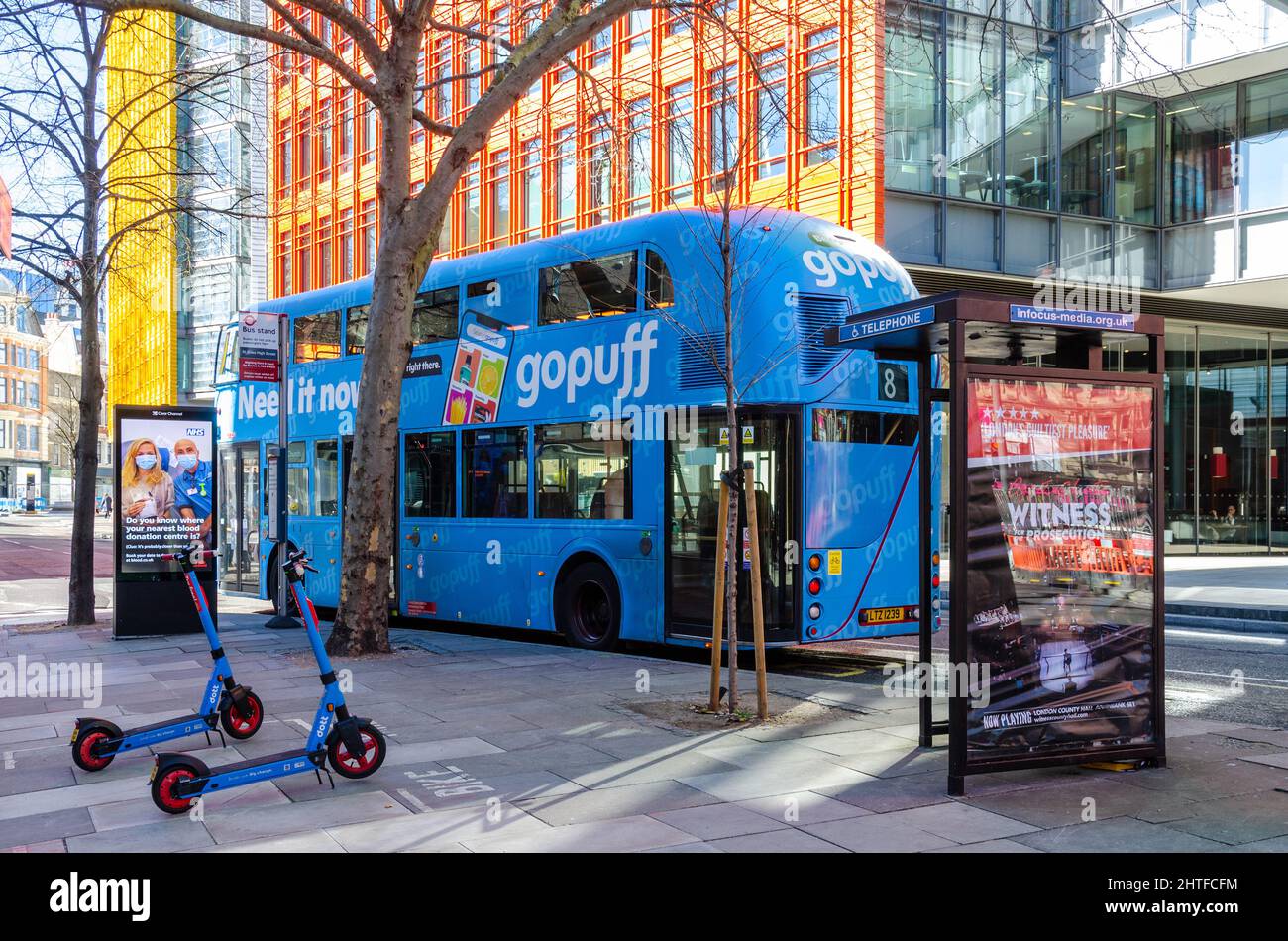 Scooters eléctricos de alquiler azules estacionados en un pavimento junto a un autobús azul de Londres frente a modernos edificios de oficinas en el centro de Saint Giles en Londres Foto de stock