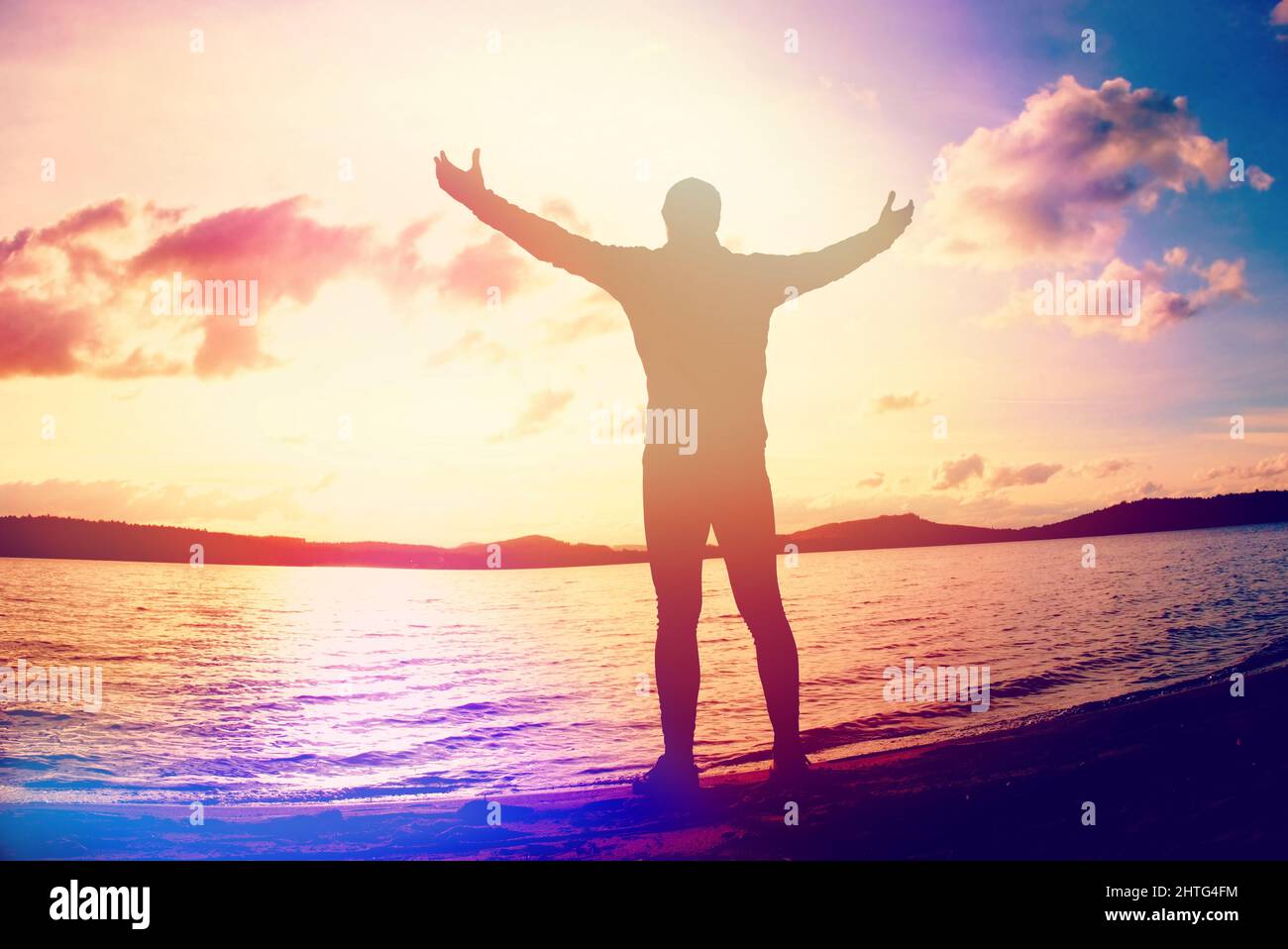 Hombre levantando los brazos en la orilla del mar. Un hombre feliz se arriesgaba con las manos por encima de la cabeza en la playa. Iluminación abstracta, llamaradas coloridas. Foto de stock