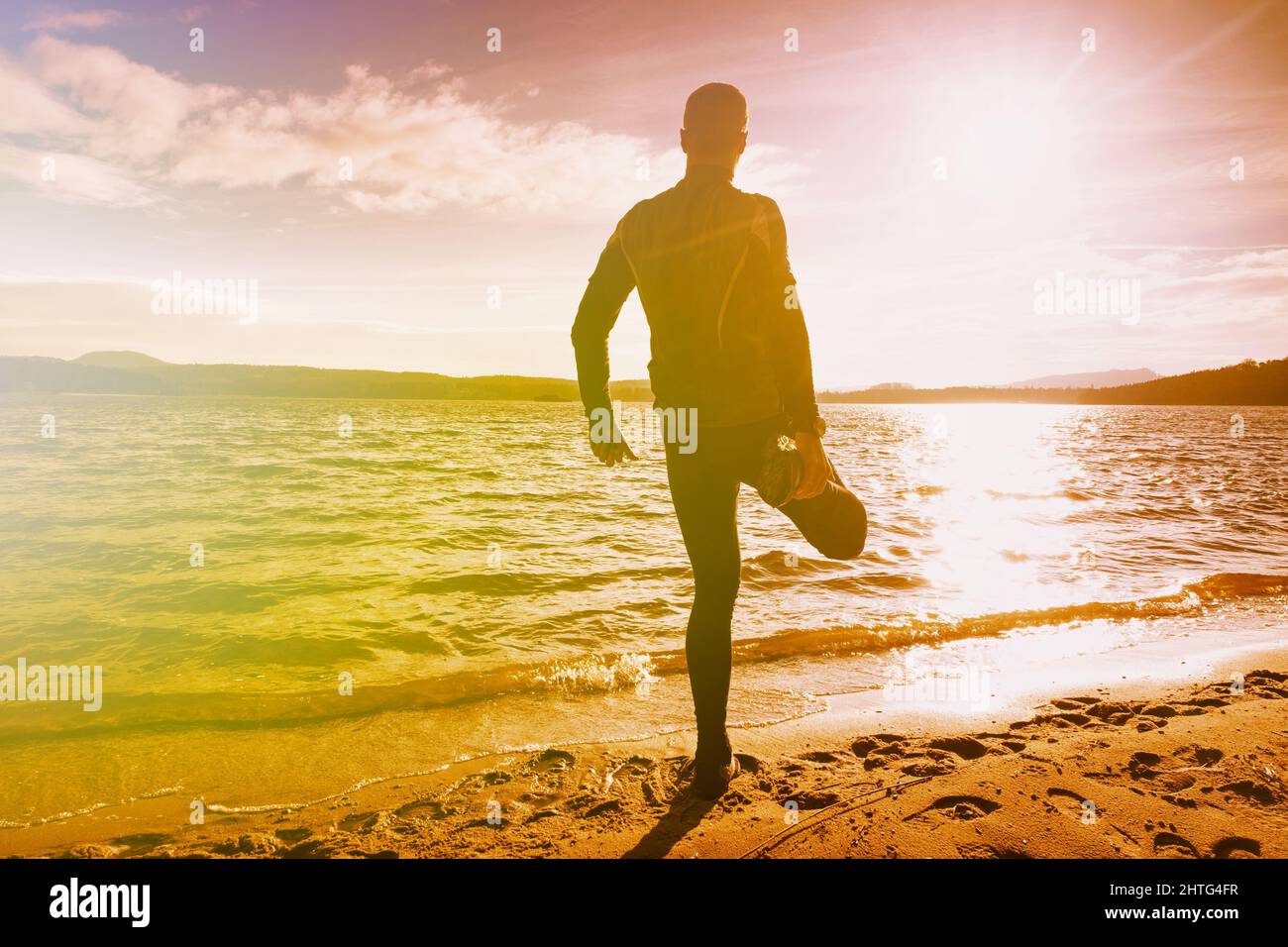 Corredor que estira la rodilla y la pierna. Ejercicios del hombre en el lago. Iluminación abstracta, llamaradas coloridas. Foto de stock