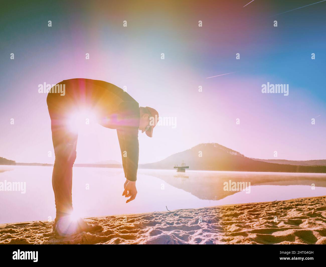 Hombre deportivo estirando se inclina hacia atrás en la playa. Corredor haciendo ejercicio en el lago. Iluminación abstracta, llamaradas coloridas. Foto de stock