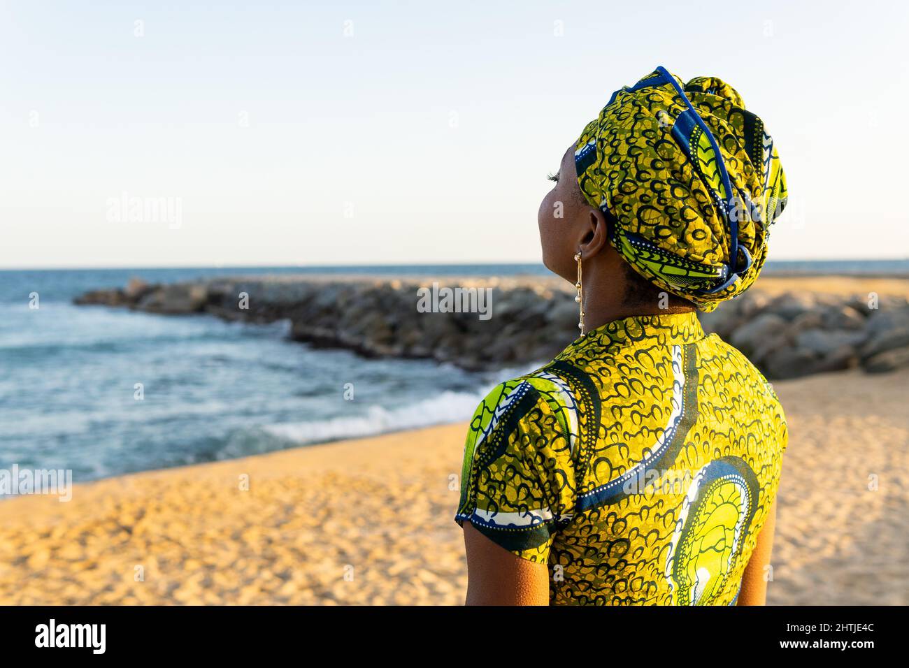 Vista posterior mujer africana en el tradicional pañuelo con la cabeza  disfrutando de la luz del sol mientras se relaja en la playa de arena  contra el mar Fotografía de stock -