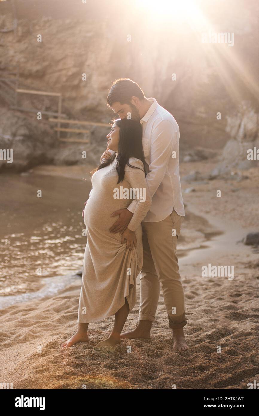 Pareja embarazada disfrutando de la puesta de sol en la playa. Mujeres embarazadas mirando el horizonte mientras su marido la besa. Foto de stock